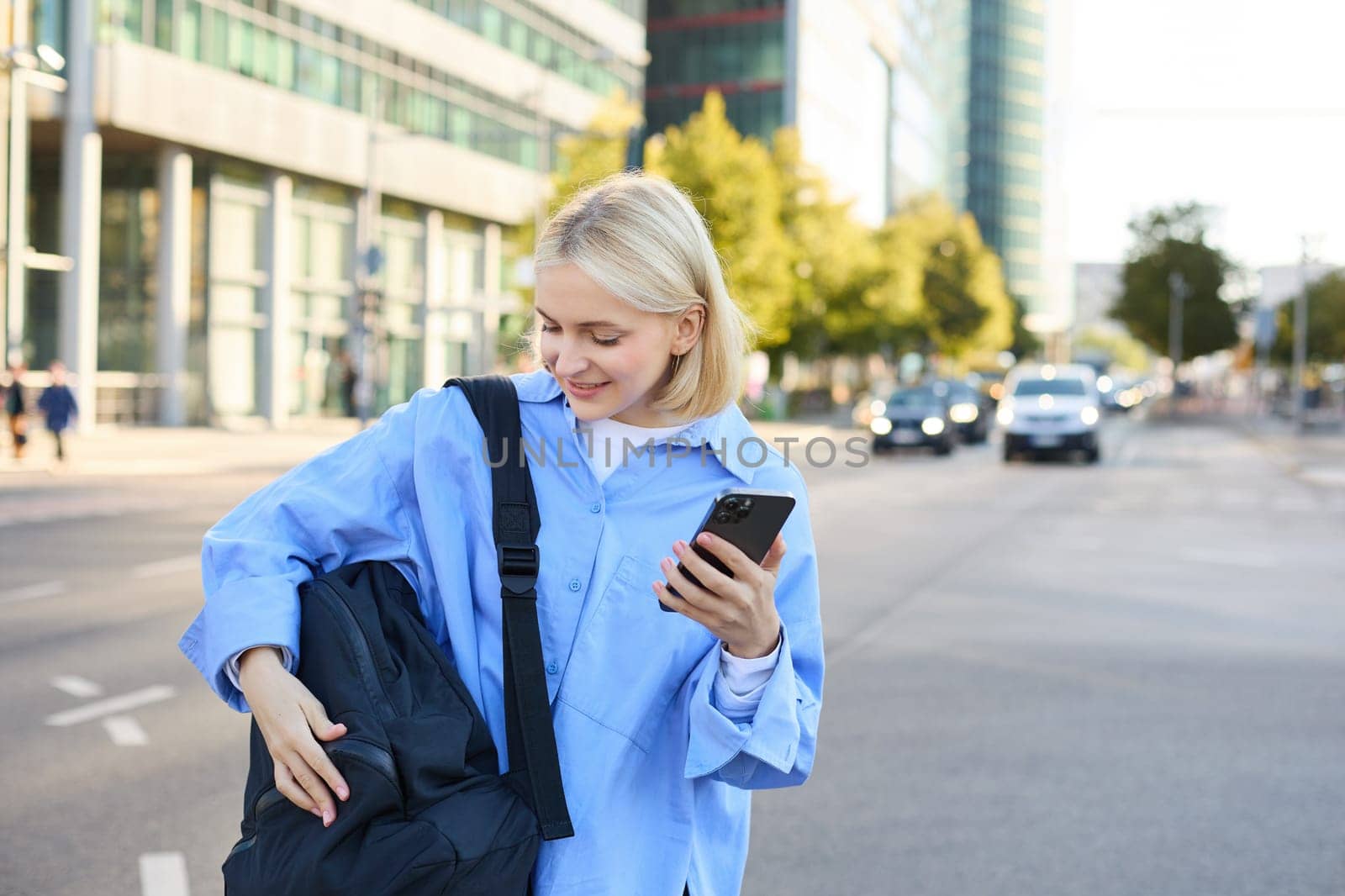 Lifestyle shot of young woman, student standing with smartphone on the street, opening her backpack pocket by Benzoix