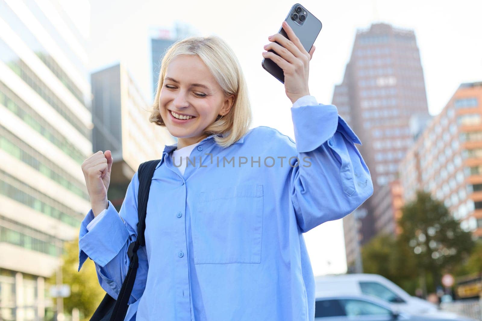 Portrait of happy, enthusiastic young woman, holding backpack and mobile phone, standing on street and rejoicing, triumphing, making fist pump gestures, dancing from happiness and joy by Benzoix