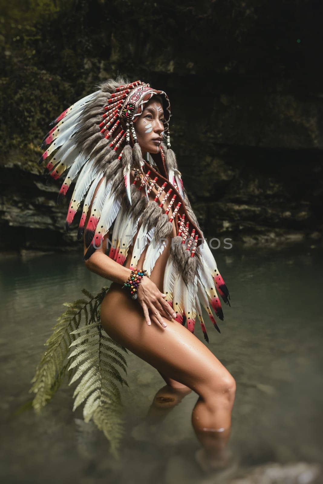 Naked girl in Native American headdresses poses sexually against the backdrop of wildlife with a fern