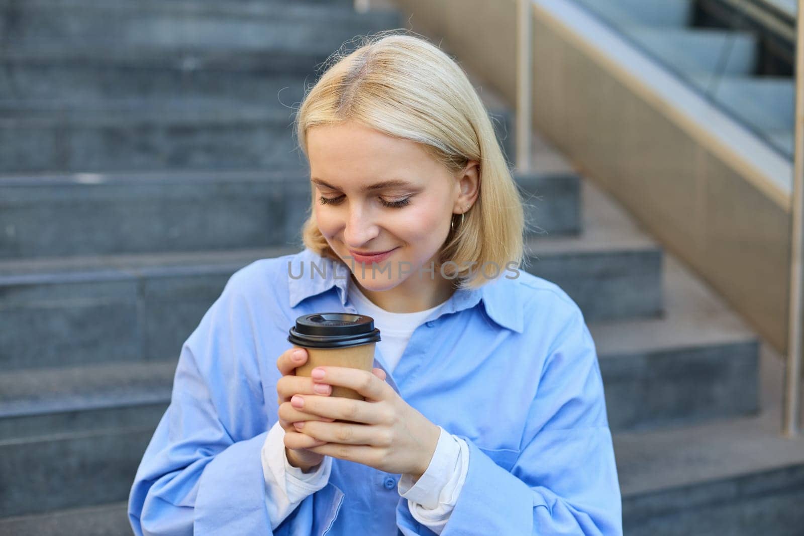 Close up portrait of beautiful, smiling blonde woman, student sitting on stairs outside campus, drinking takeaway coffee, warm-up her hands while holding a cup.