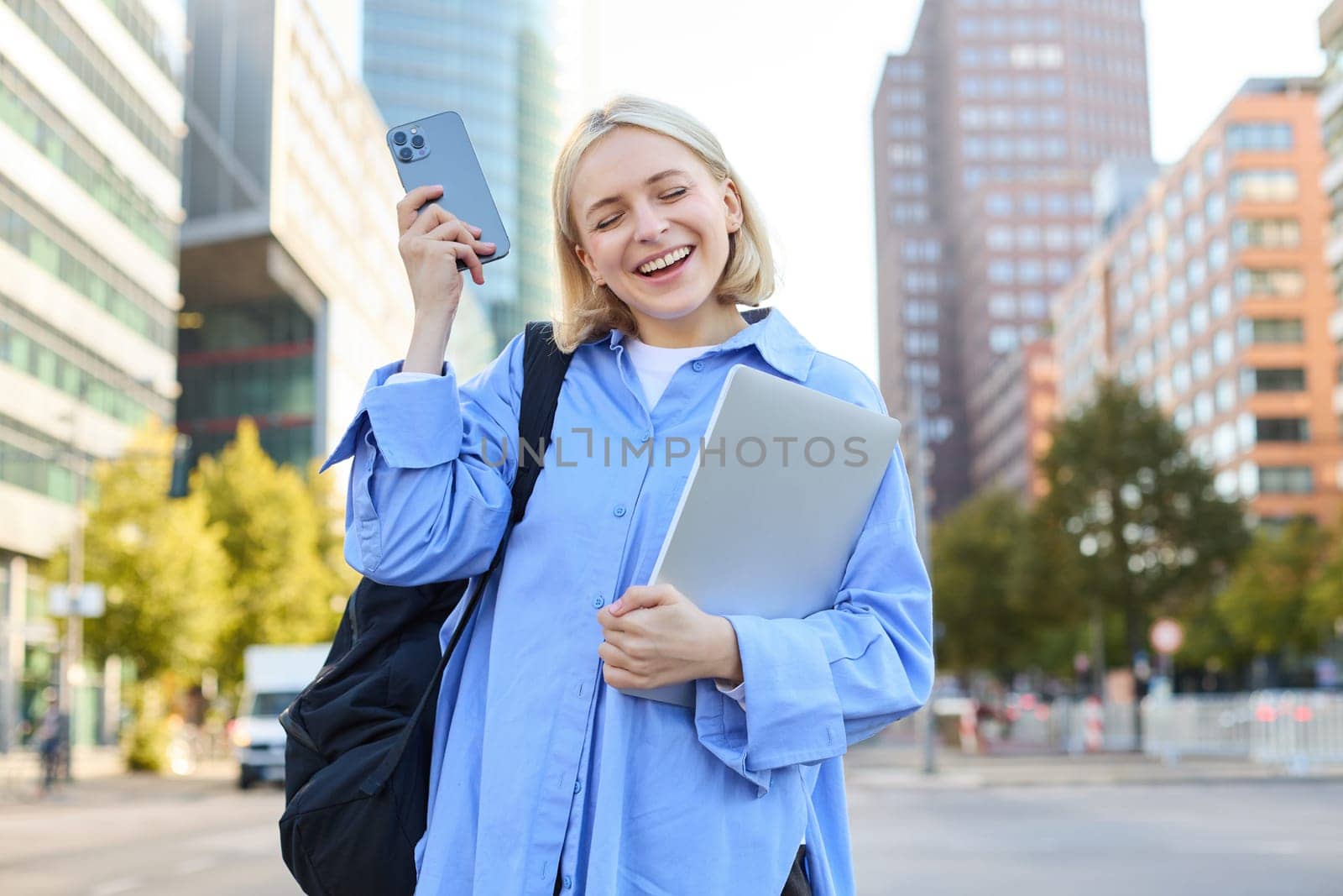 Enthusiastic blond woman, shaking smartphone in hand, holding laptop, standing on street of city centre, laughing and smiling, looking pleased, triumphing.