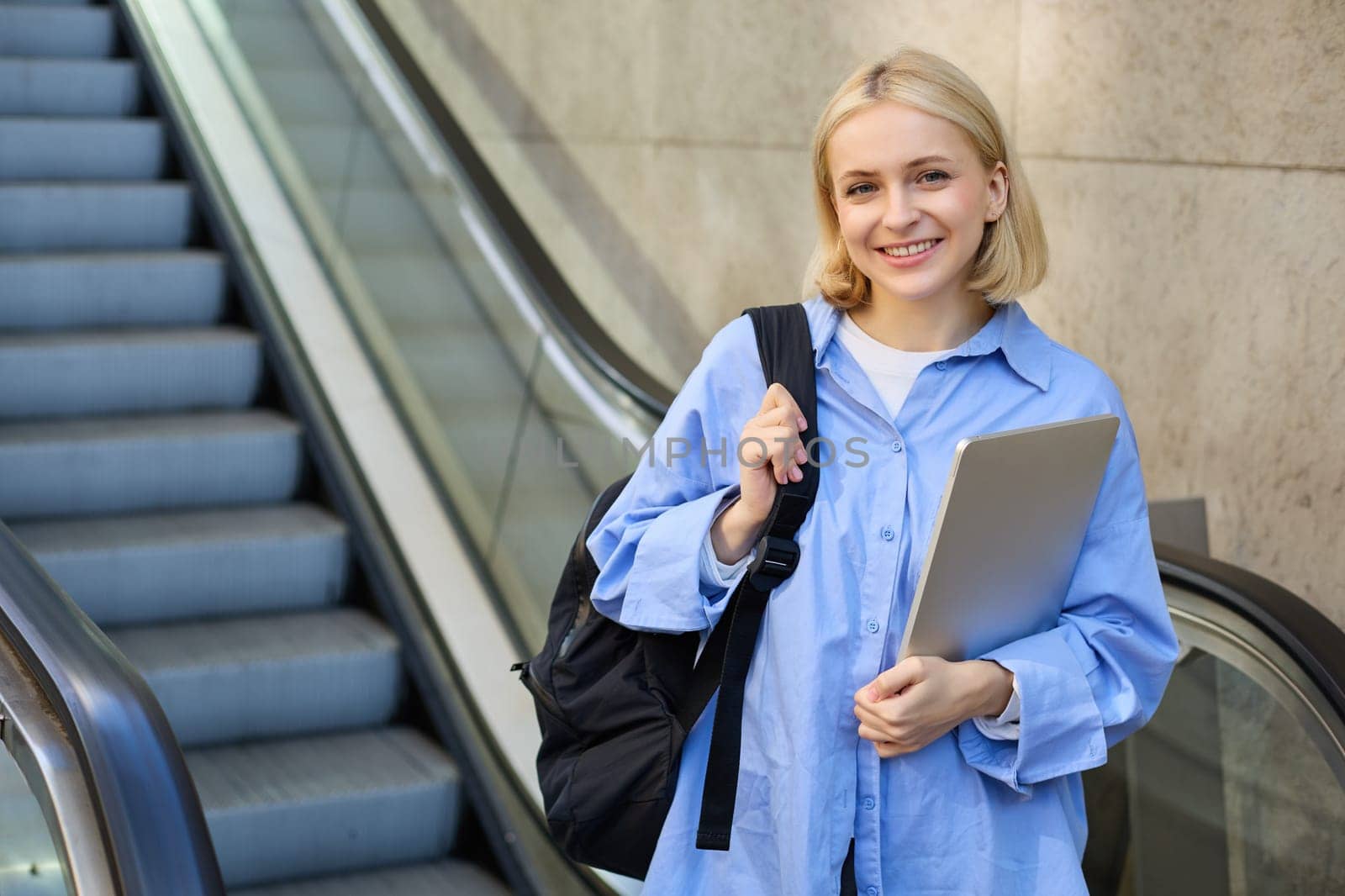 Concept of education and people. Young woman with backpack, carries laptop in hand, standing near escalator, going to tube, on her way to work on university by Benzoix