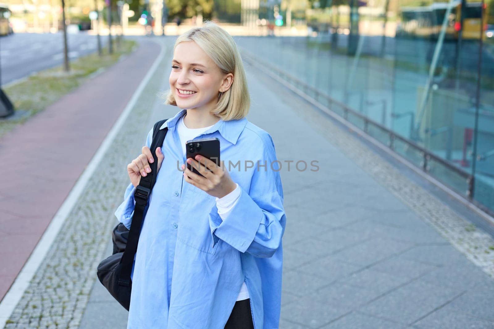 Portrait of smiling, modern blond woman walking along the street in blue collar shirt, has backpack on shoulder, messaging, using mobile phone app.