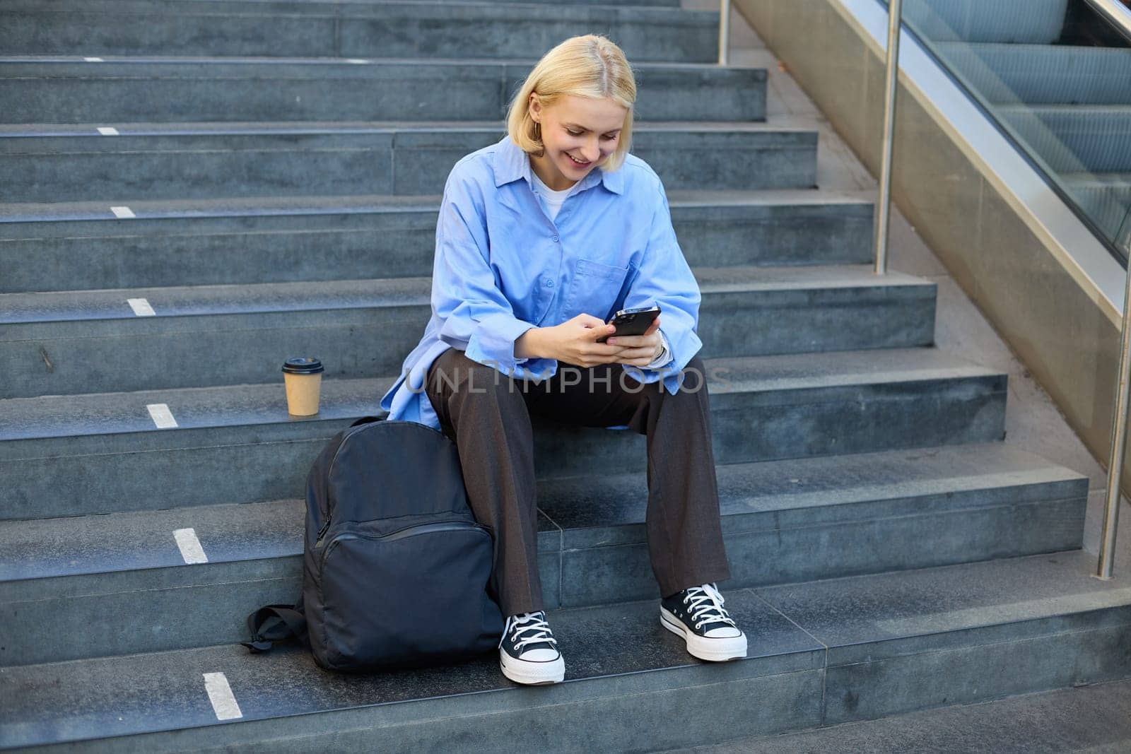 Portrait of young modern woman, student with backpack, sitting on city stairs on lunch break, using smartphone, looking aside by Benzoix