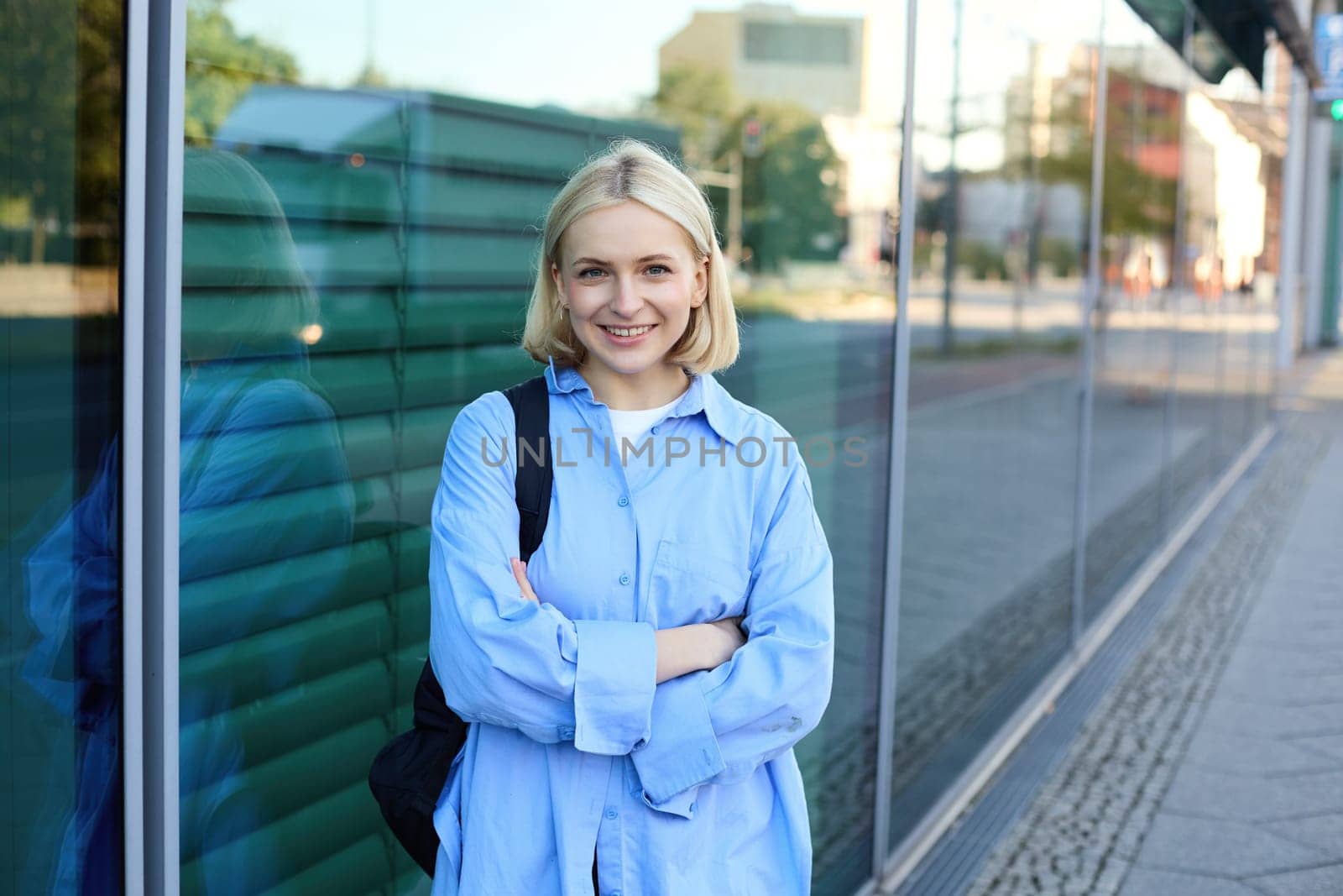 Image of young modern woman on street, college student in blue shirt, with backpack, cross arms on chest and smiles with confidence at camera.