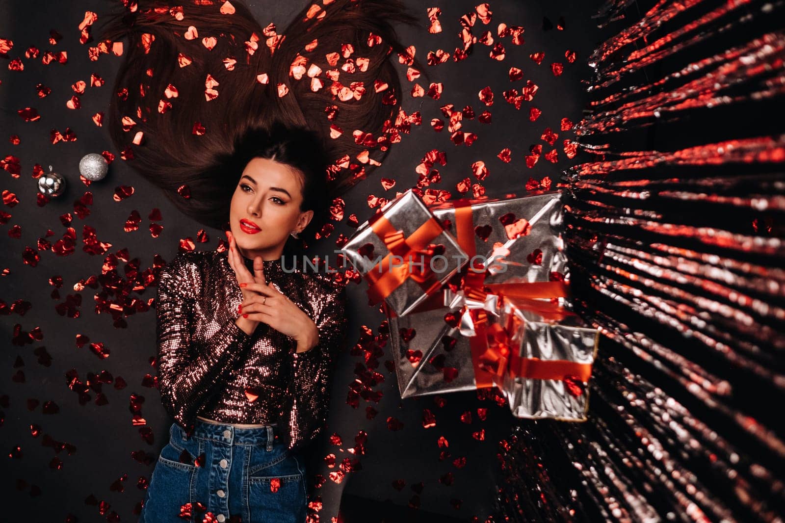 Top view of a girl lying in shiny clothes on the floor in confetti in the form of hearts and gifts by Lobachad