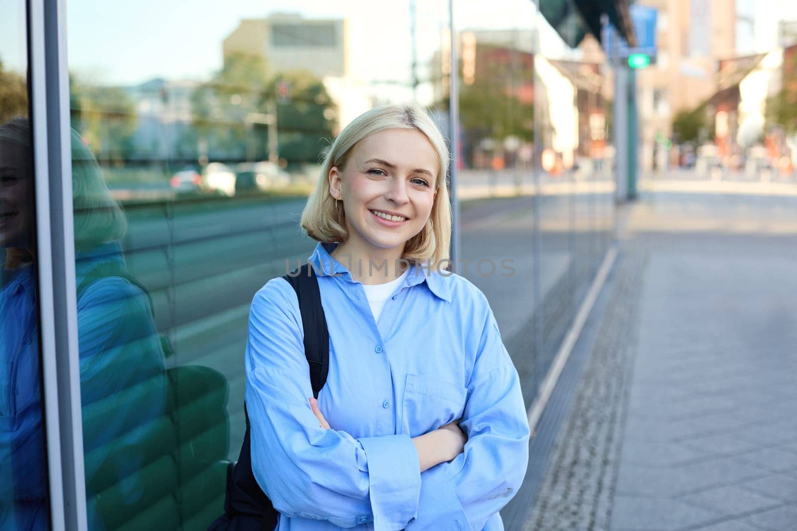 Lifestyle portrait of young smiling woman, student or office employee, standing on street in blue shirt, cross arms on chest and looking confident at camera by Benzoix