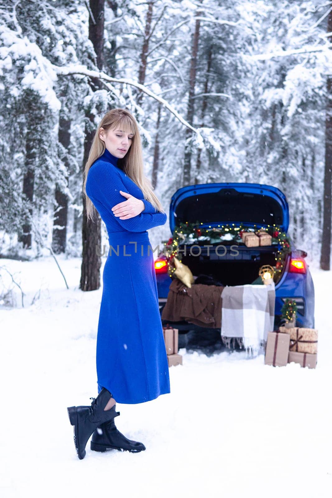 Woman in winter snowy forest in blue dress next to blue car decorated with Christmas decor. Christmas and winter holidays concept