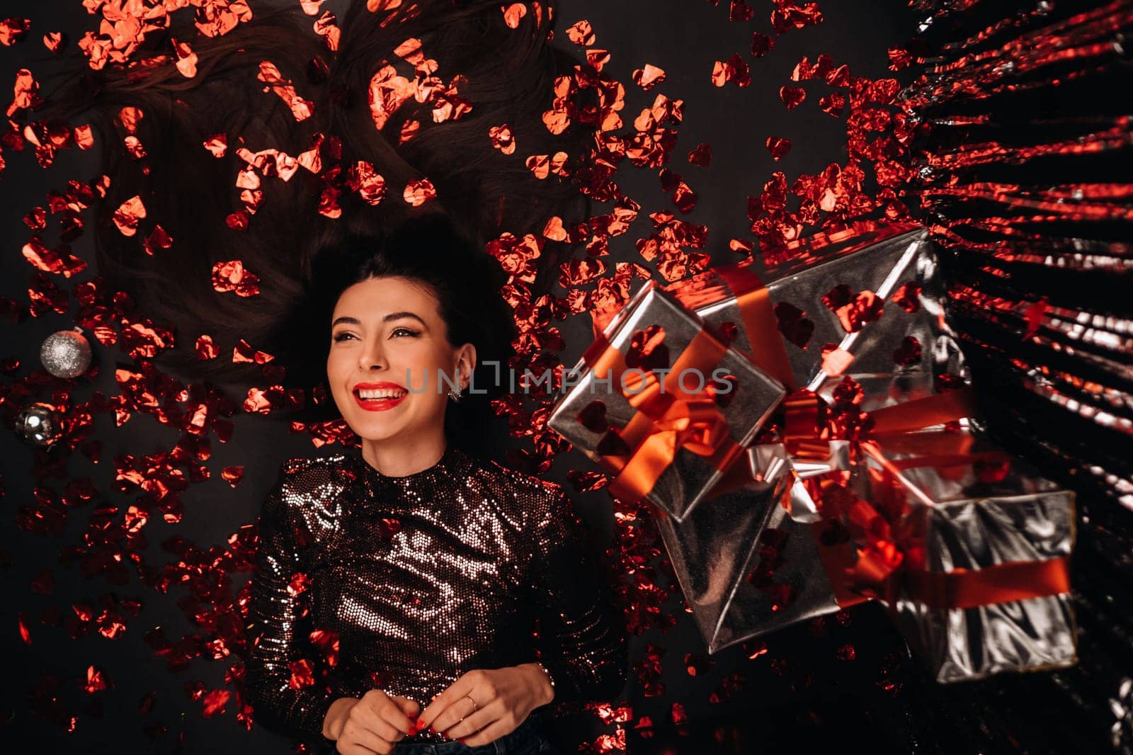 Top view of a girl lying in shiny clothes on the floor in confetti in the form of hearts and gifts.