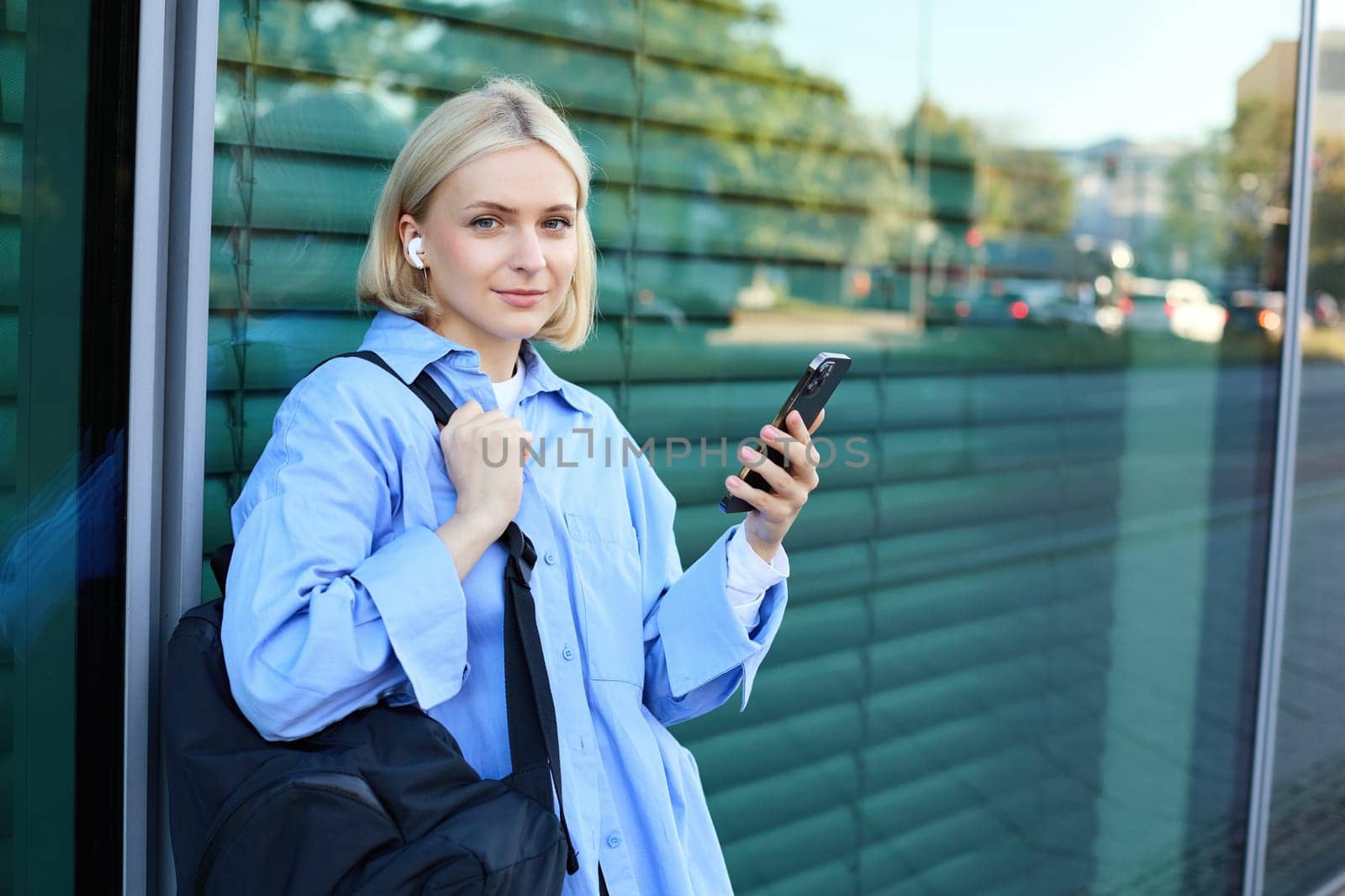 Lifestyle portrait of smiling young female model, student with backpack, waiting for someone on street, standing outdoors with smartphone by Benzoix