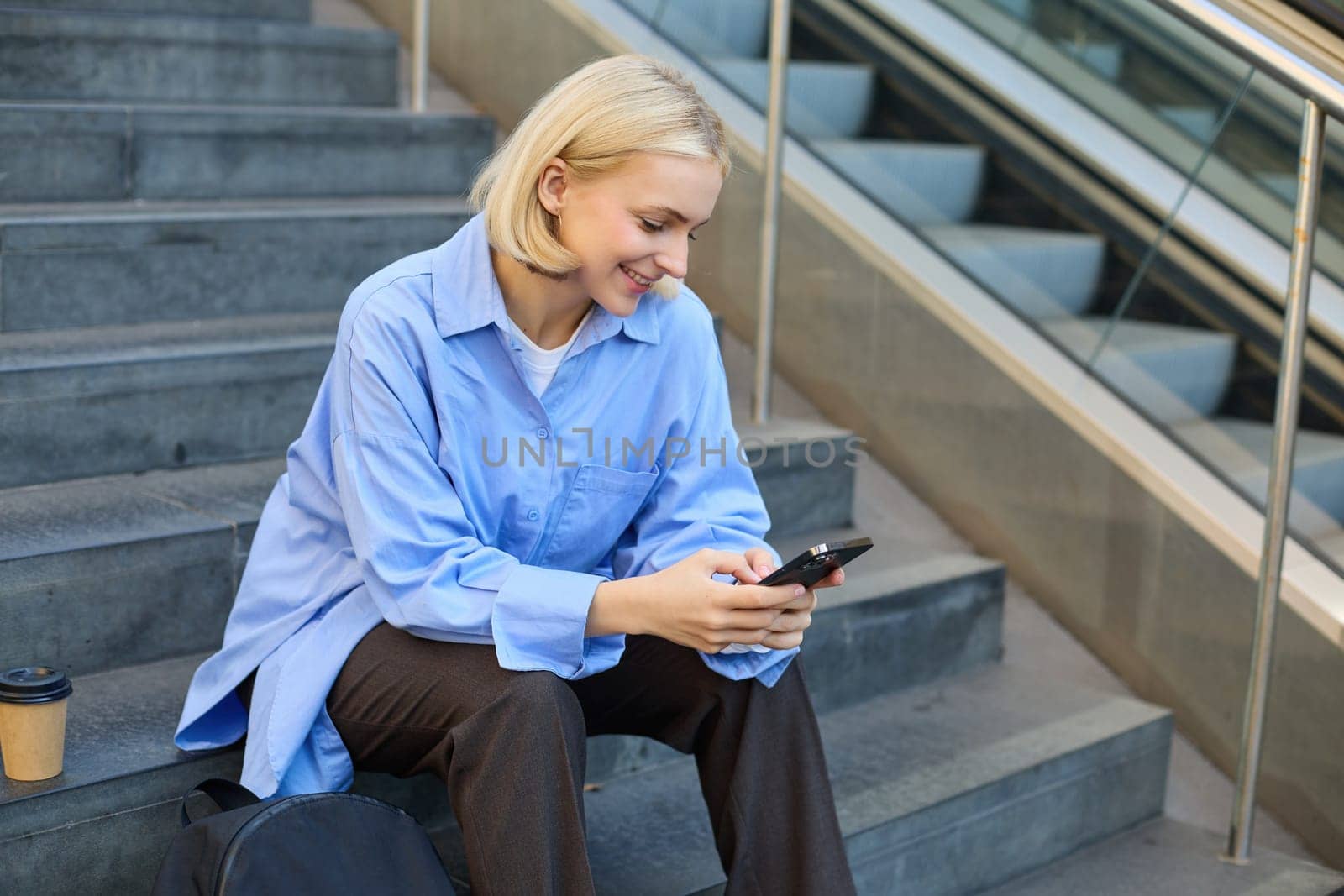 Lifestyle portrait of smiling, young woman, sitting on street stairs, looking at mobile phone, reading a message on smartphone, drinking coffee on lunch break by Benzoix