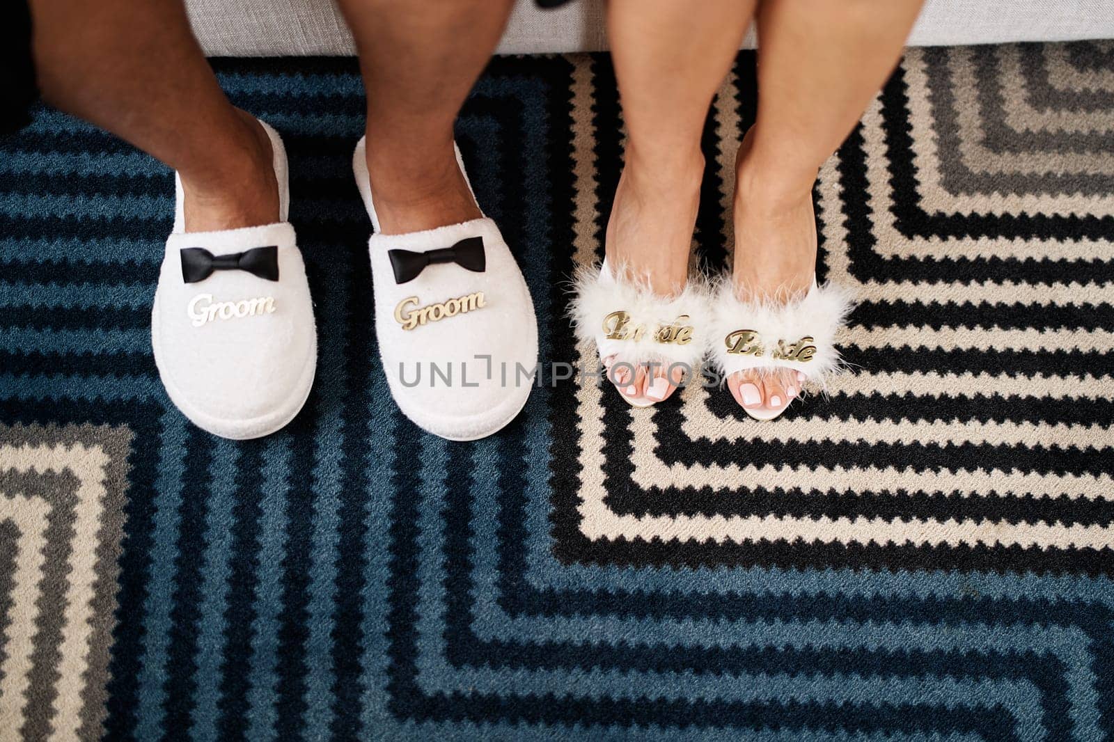 Male and female feet in slippers stand on a striped carpet. Caption: Groom. Bride. Cropped, faceless by Nadtochiy