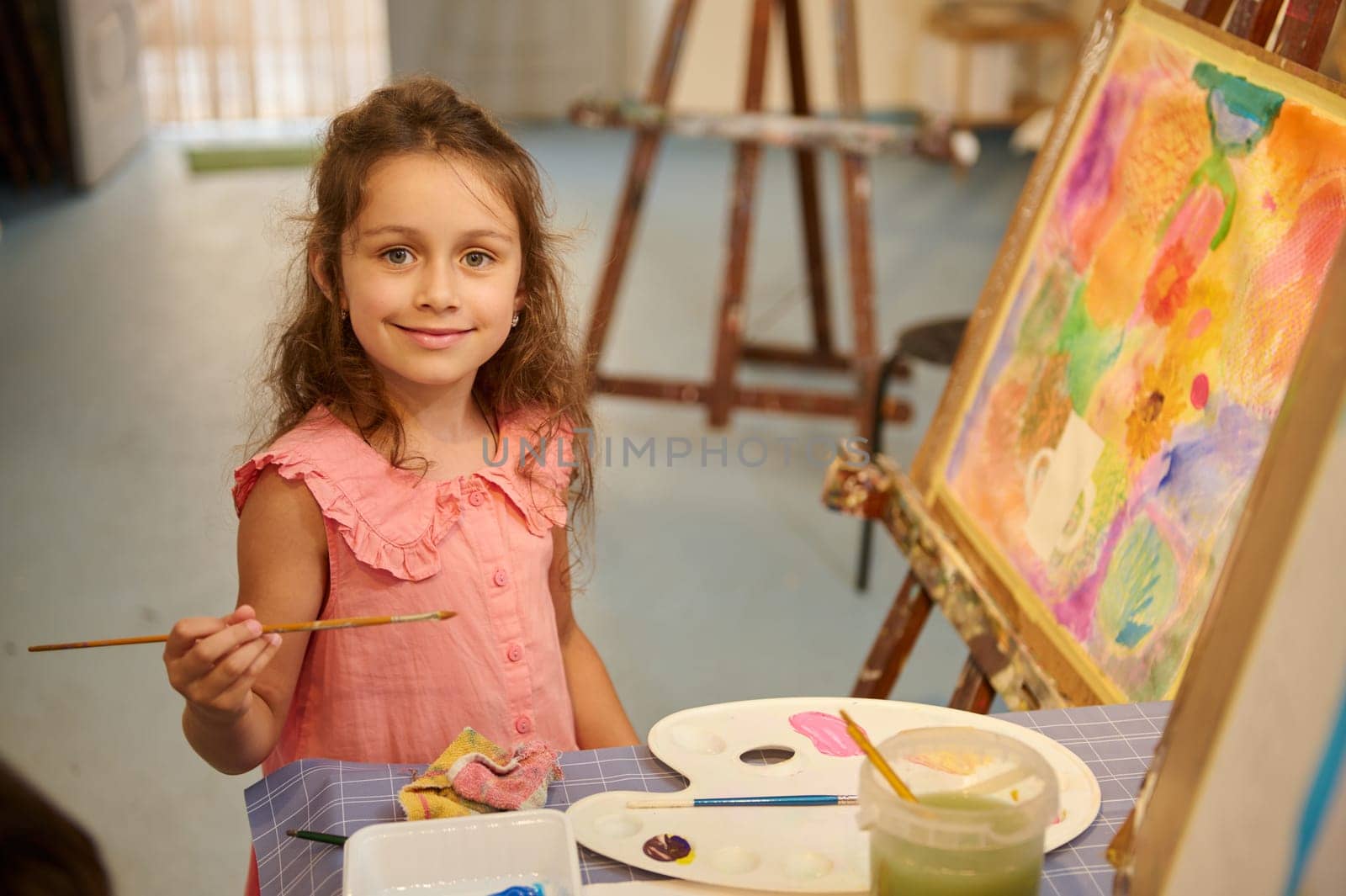 Adorable little child girl artist smiling cutely looking at the camera, sitting near picture indoor while learning fine art in a creative studio. Kids. Education. Learning drawing and painting.