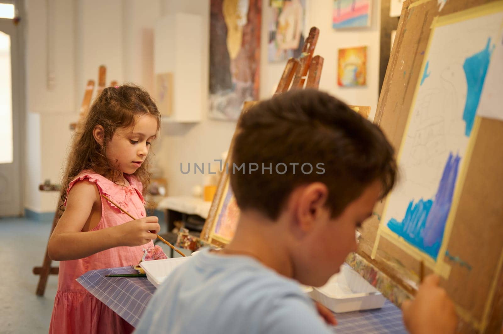 Adorable talented kids, little artists standing in front of easel, holding brush and palette, drawing in fine art studio by artgf