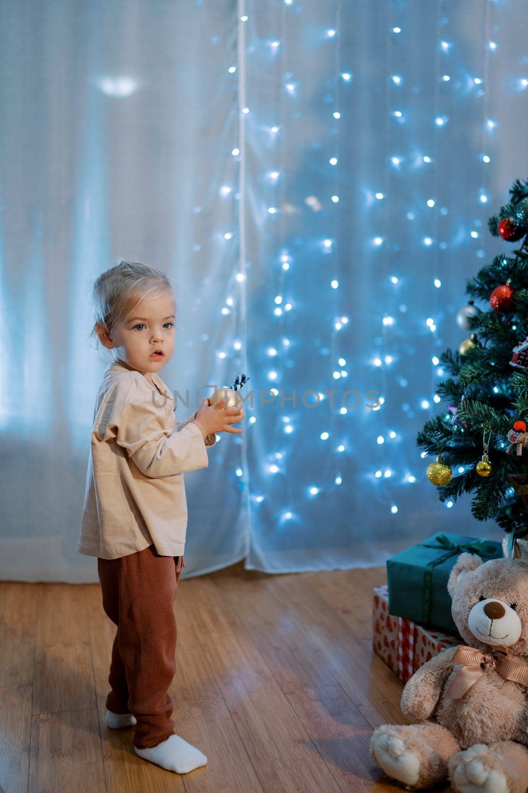 Little girl stands near a decorated Christmas tree with a ball in her hands. High quality photo