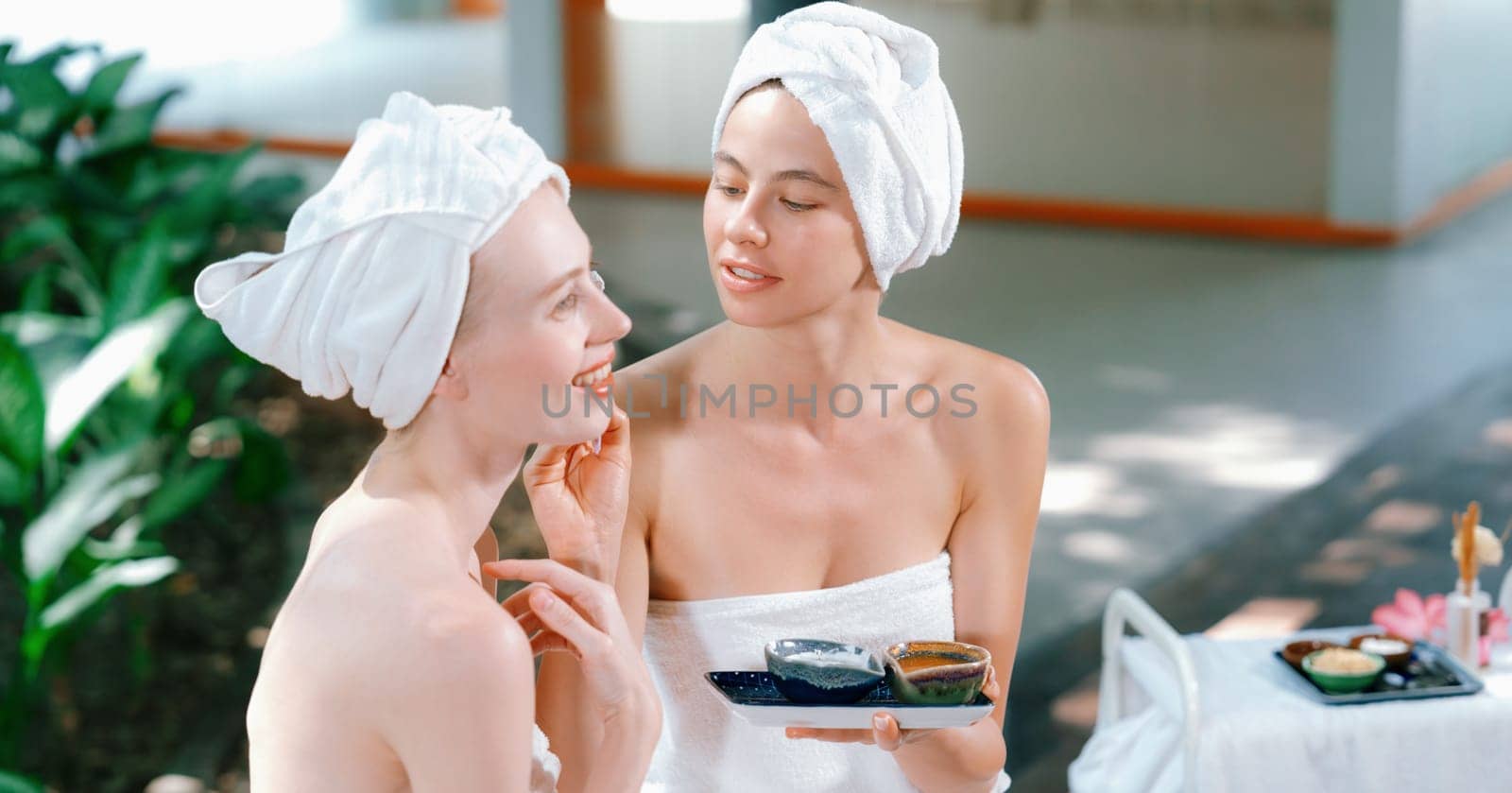 A portrait of beautiful woman in white towel receiving herbal facial mask by her friend at outdoor. Attractive spa girl putting face mask on pretty female with beautiful skin. Tranquility.