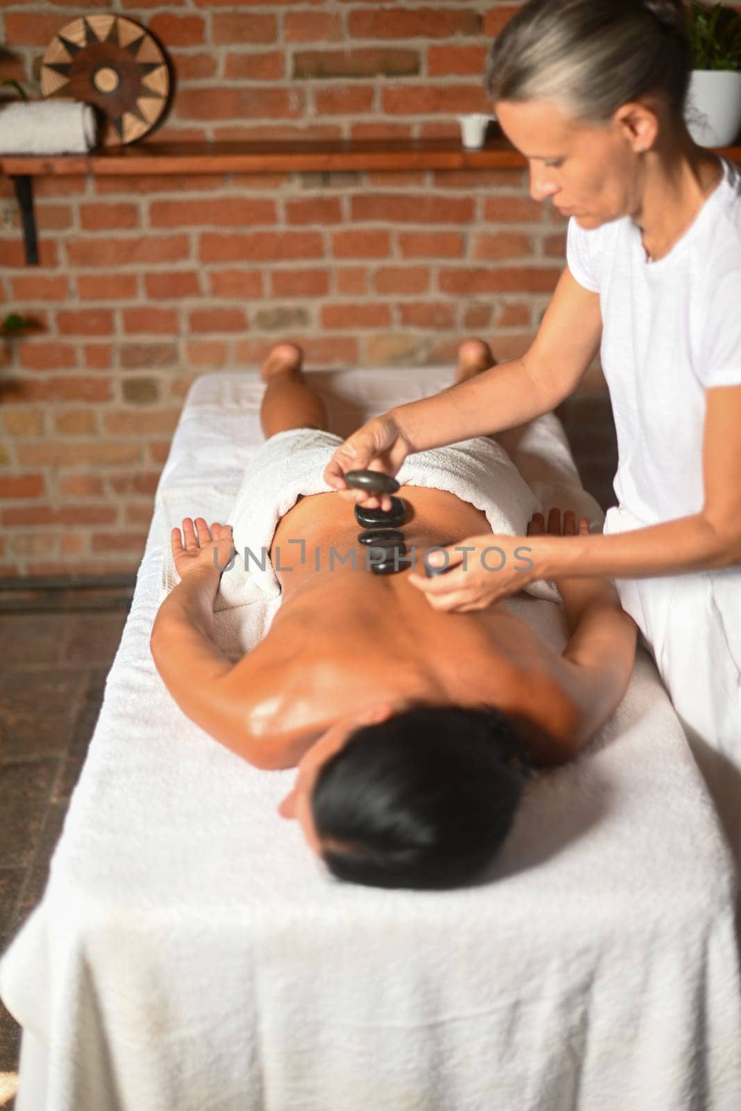 professional caucasian female physiotherapist masseuse performing stone massage on backbone to hispanic latino brunette woman in spa clinic