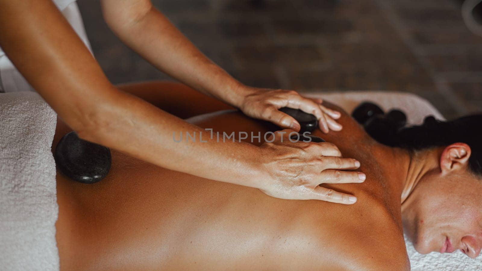 professional caucasian female physiotherapist masseuse performing stone massage on backbone to hispanic latino brunette woman in spa clinic