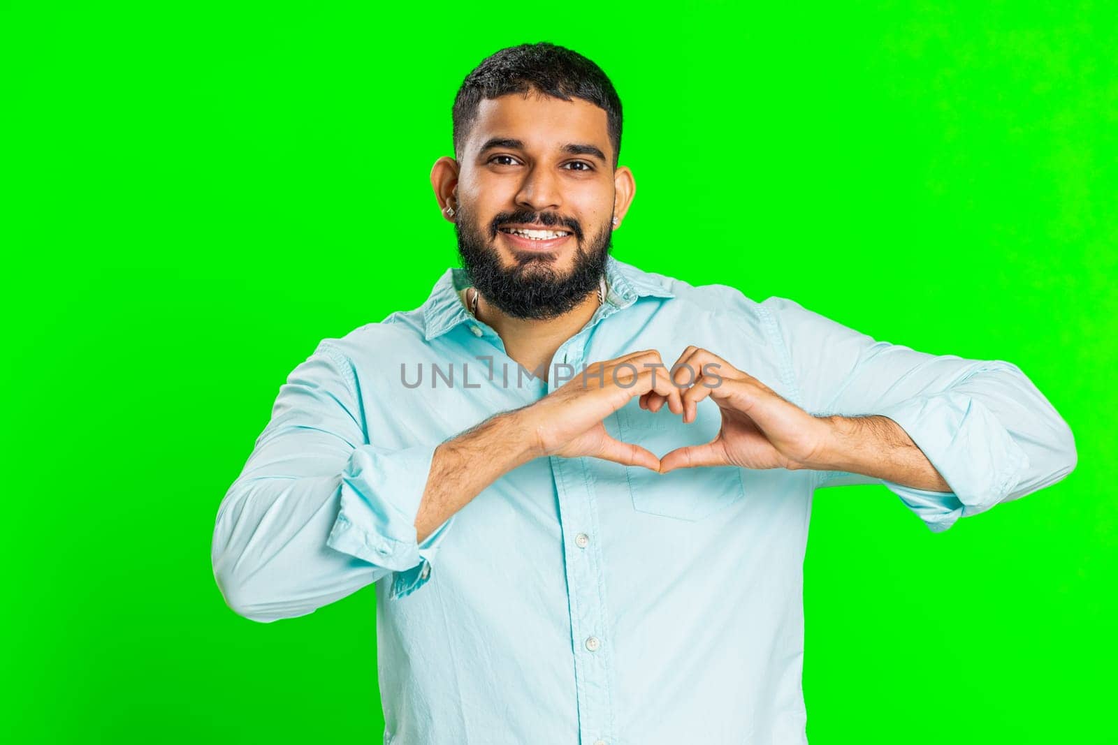 Man in love. Smiling attractive Indian man makes heart gesture demonstrates love sign expresses good positive feelings and sympathy. Handsome Arabian young guy isolated on chroma key wall background