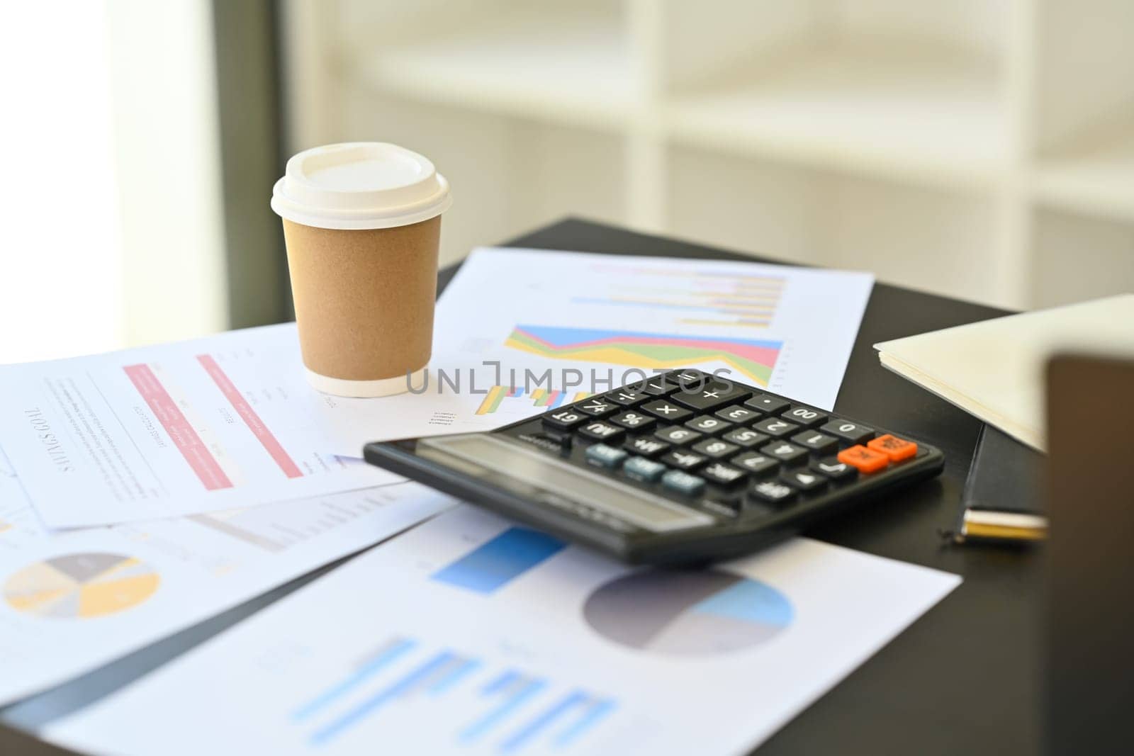Financial analyst workplace with calculator machine, notepad, documents and paper cup on table by prathanchorruangsak
