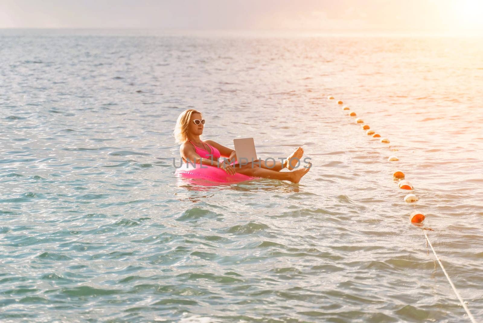 Woman works on laptop in sea. Freelancer, blond woman in sunglases floating on an inflatable big pink donut with a laptop in the sea at sunset. People summer vacation rest lifestyle concept