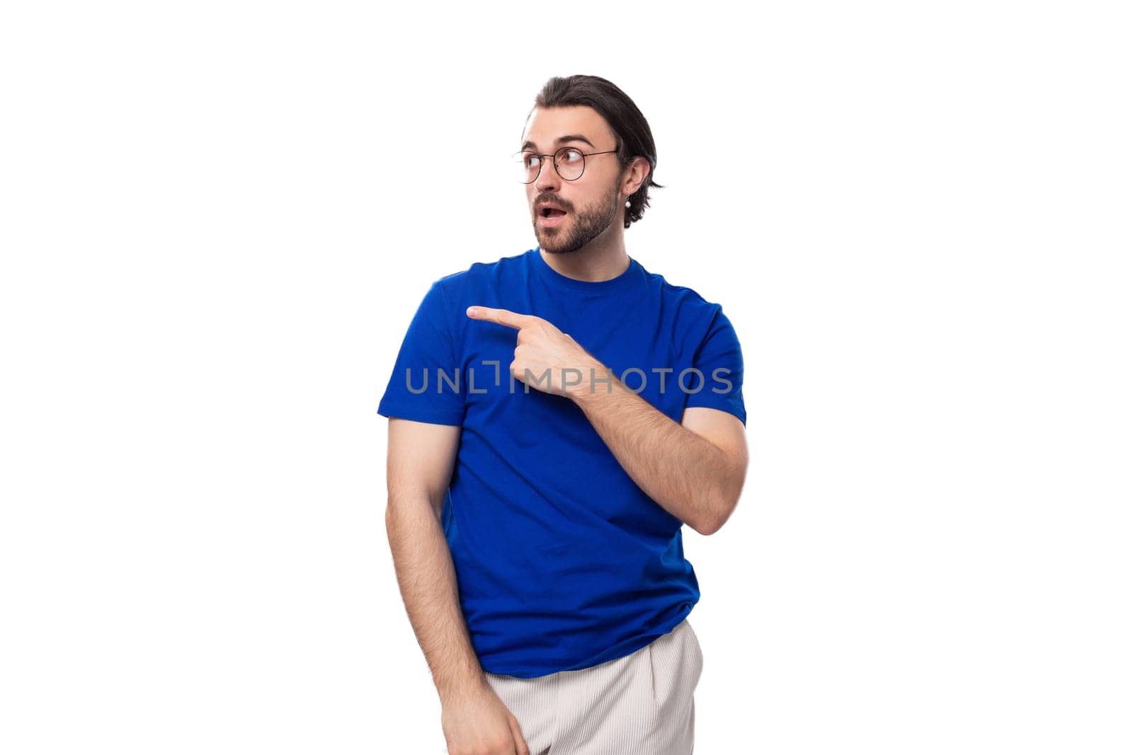 young confident brunette man with a beard dressed in a blue t-shirt shows his hand to the space for advertising on a studio white background by TRMK