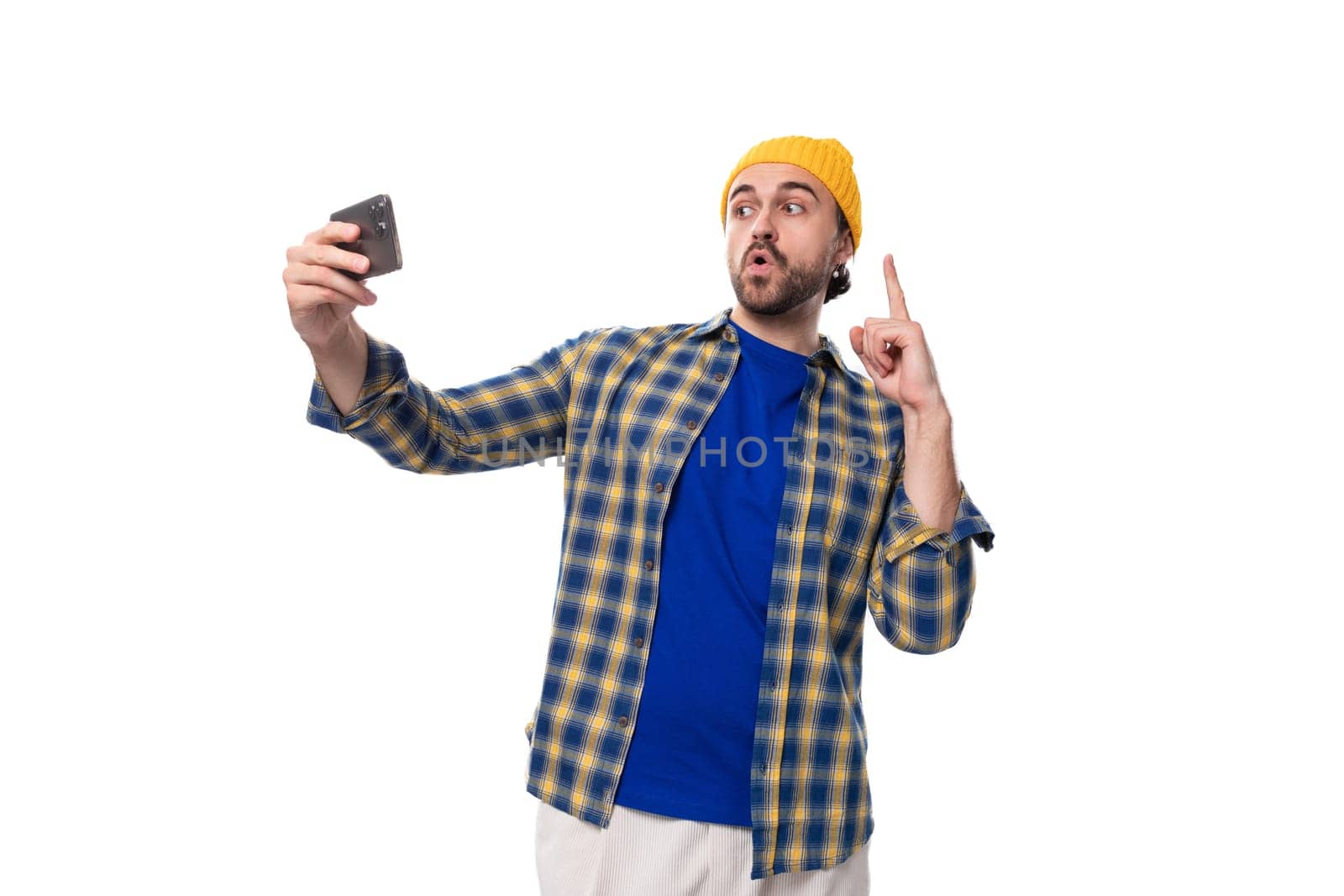 young european brunette man with a beard and mustache dressed in a yellow cap and a blue shirt takes a selfie on a white background with copy space.
