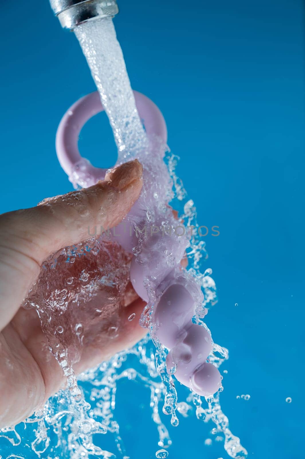 Woman holding lilac anal beads under running water on blue background. Sex toy hygiene concept