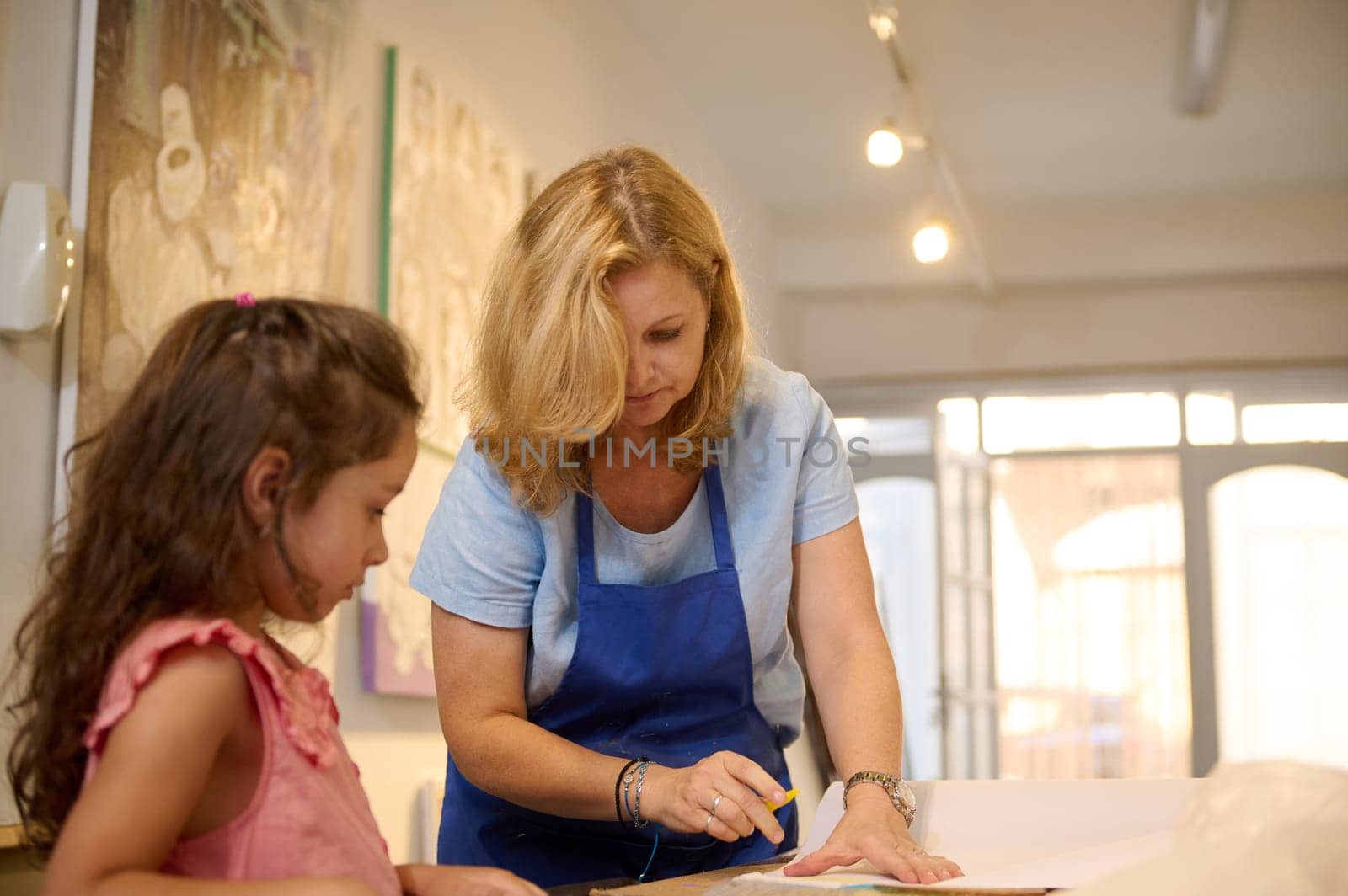 Authentic professional portrait of a female painter artist teaching paint art to a little student girl in creative workshop. Visual and fine art lesson in cozy studio. People. Creativity. Inspiration.