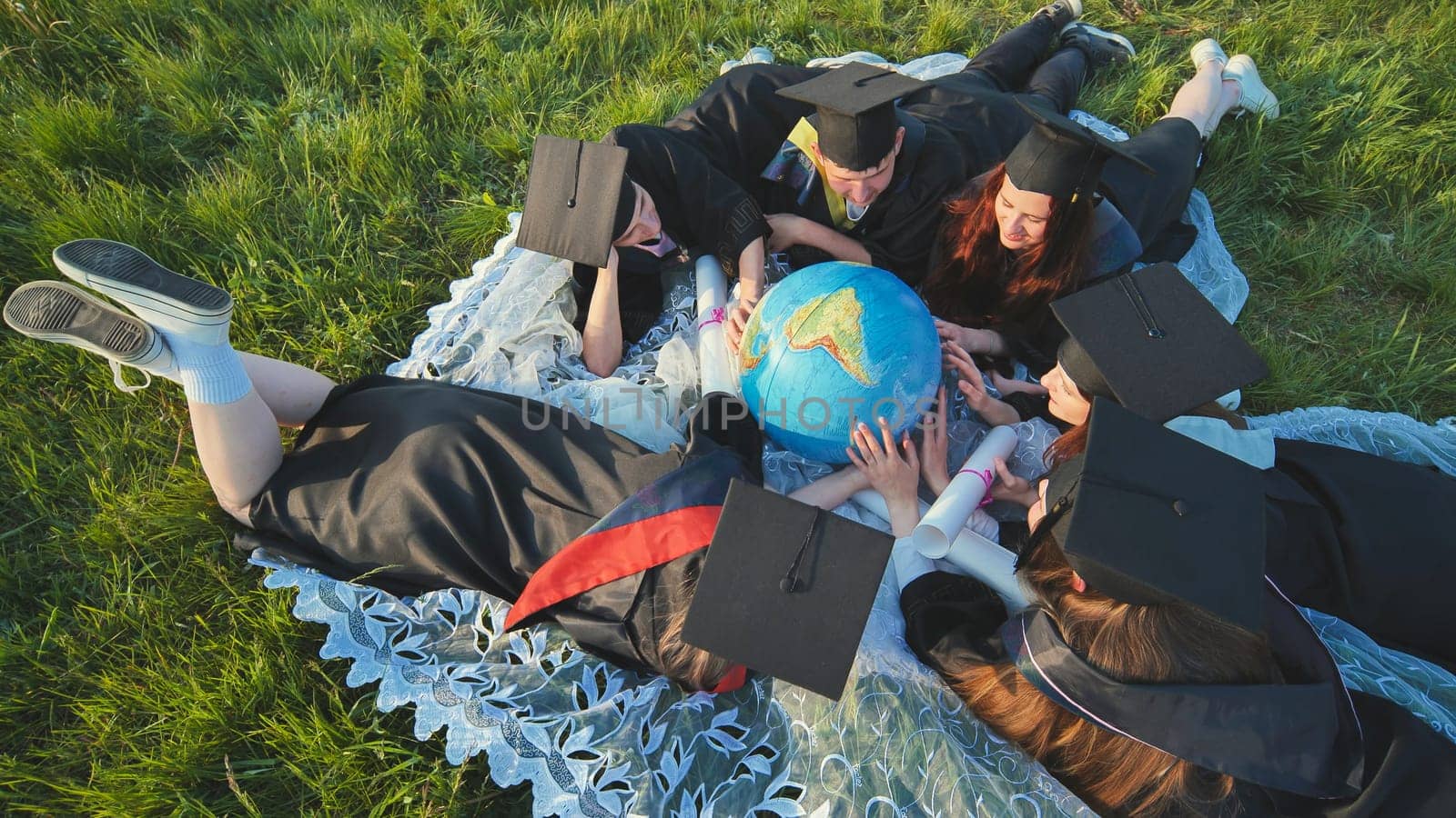 Graduate students in black robes study a globe on the grass