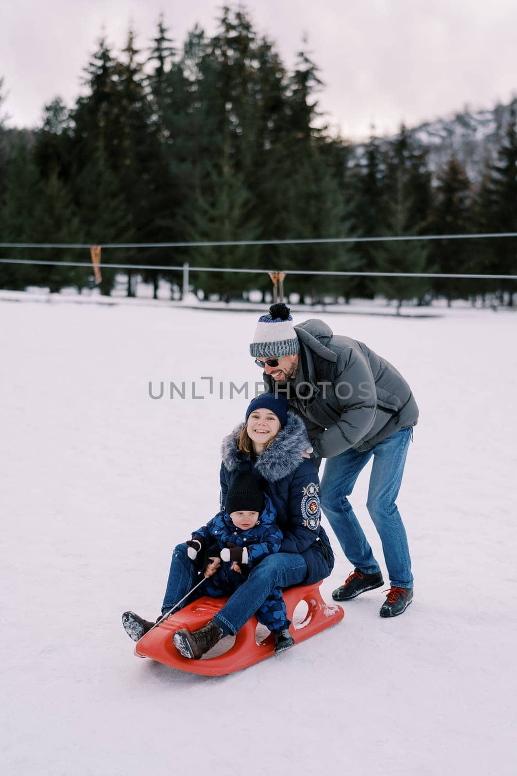 Dad pushes a smiling mom with a little boy on a sled down a snowy hill. High quality photo