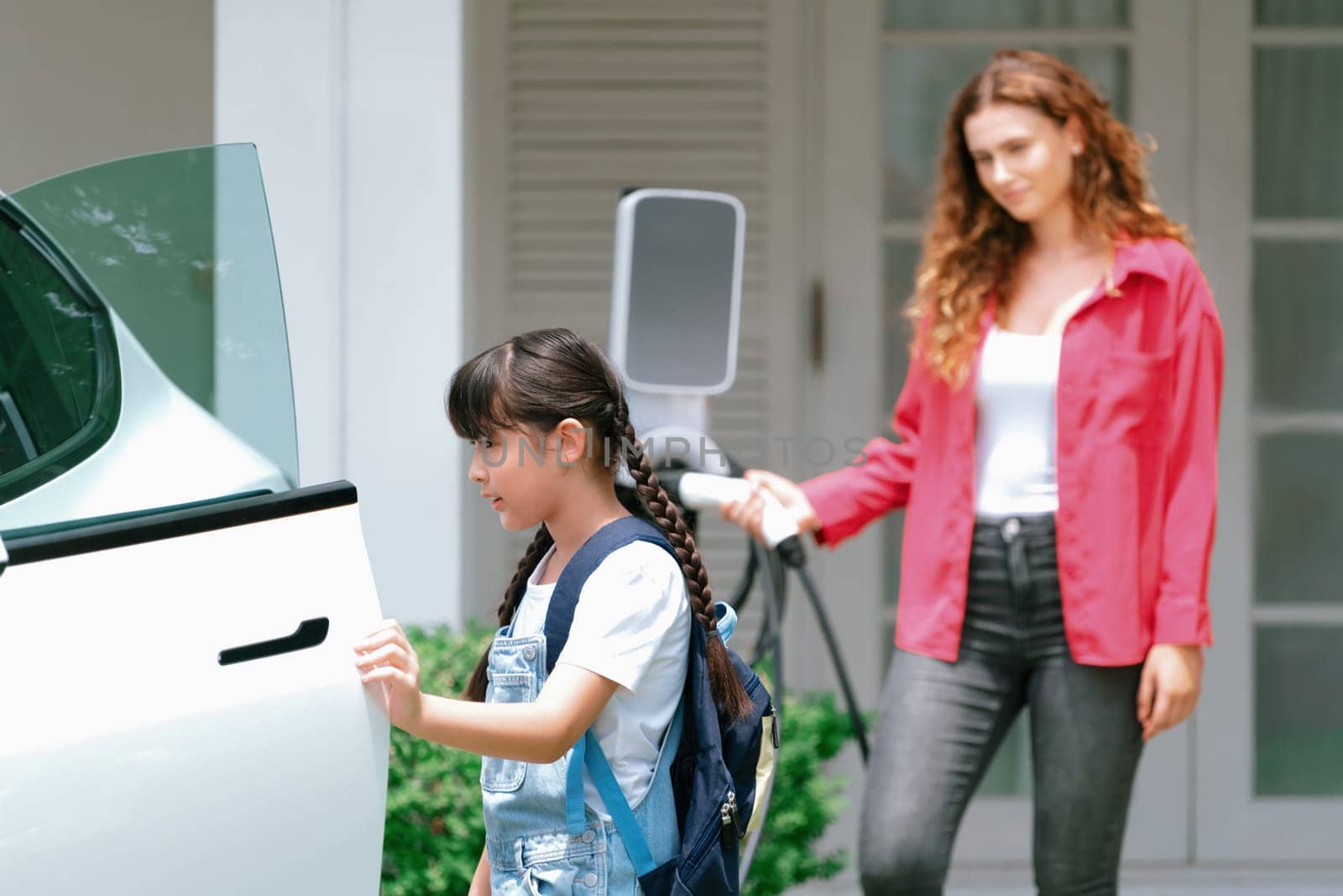 Happy little young girl learn about eco-friendly and energy sustainability as she help her mother recharge electric vehicle from home EV charging station. EV car and modern family concept. Synchronos