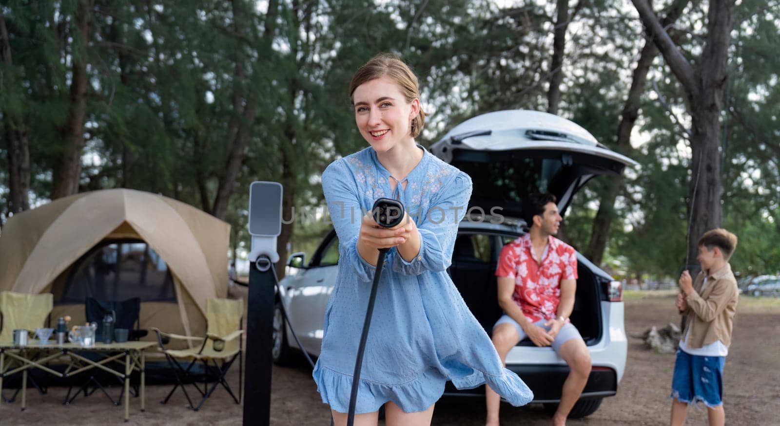 Outdoor adventure and family vacation camping at sea travel by eco friendly car. Cheerful woman or mother holding, pointing EV charger point with playful and happiness posture in campsite. Perpetual