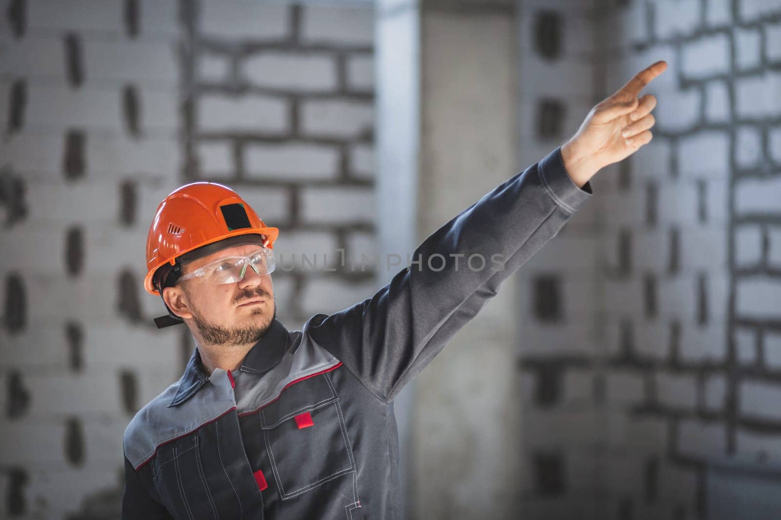 Bearded worker in blue uniform and helmet pointing with finger to the side at construction site.
