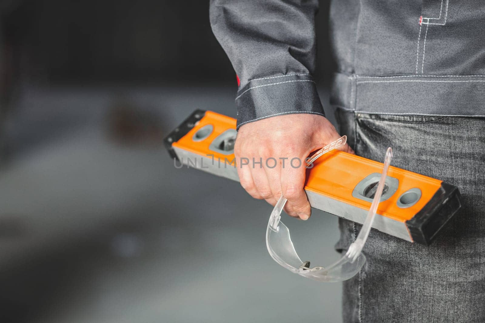 Construction level and safety glasses in the hand of a construction worker in overall. DIY or home renovation concept, copy space.