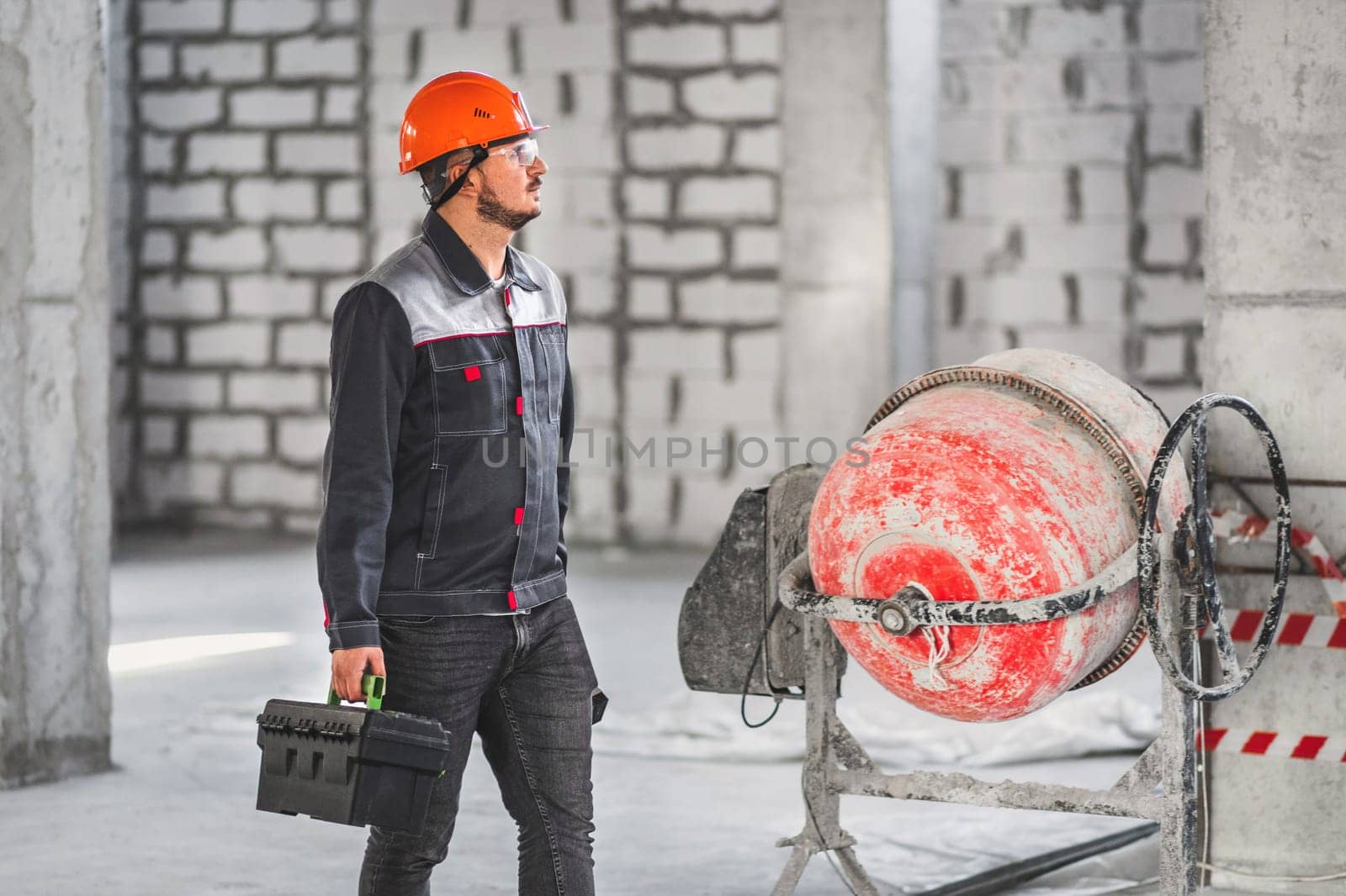 A worker with a suitcase with tools, in overalls, walks along a construction site past a concrete mixer.