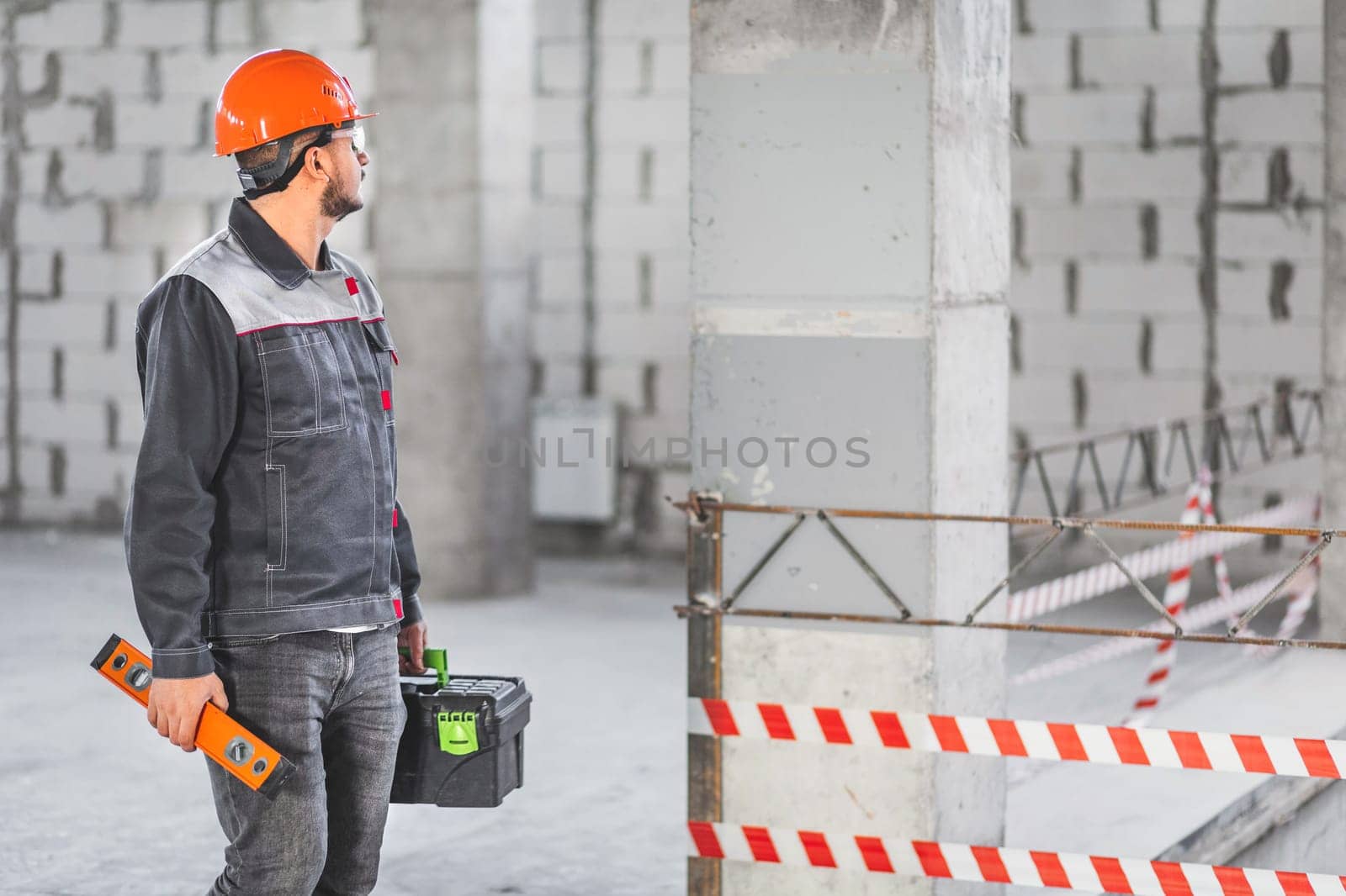 A foreman inspects a new apartment before starting work. Builder in uniform carrying toolbox and level tool at construction site, copy space by Rom4ek