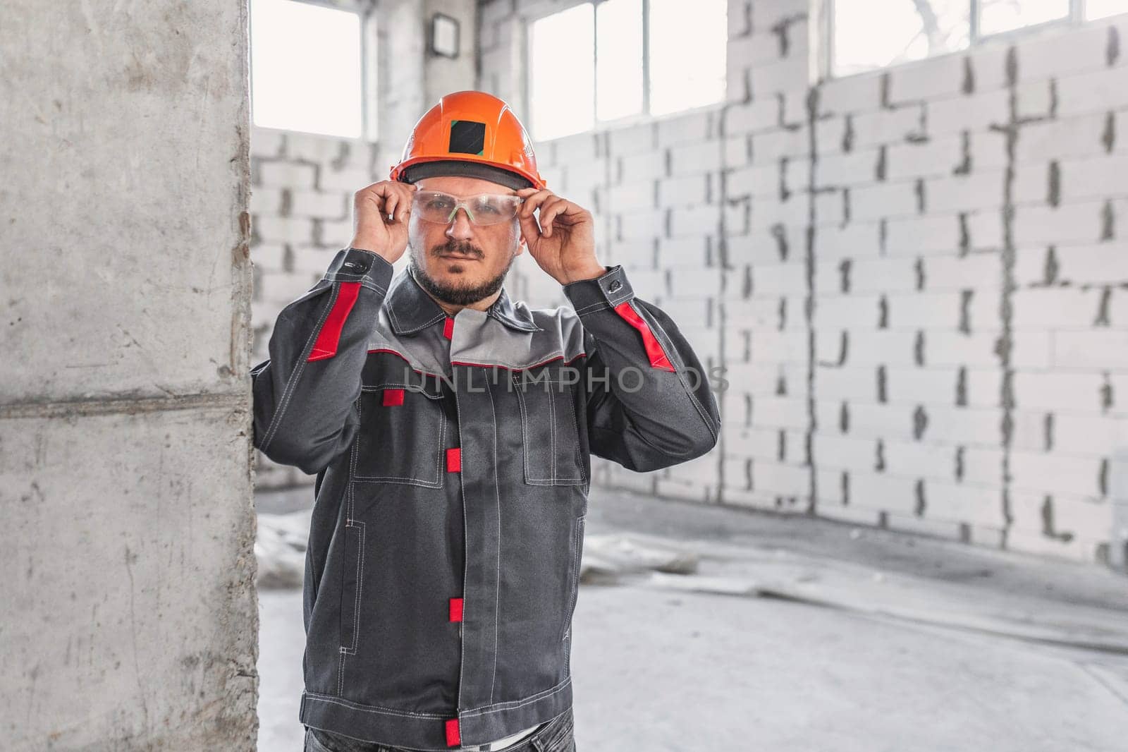 Construction worker in a hard hat and overalls adjusts his safety glasses at a construction site, workplace safety concept.