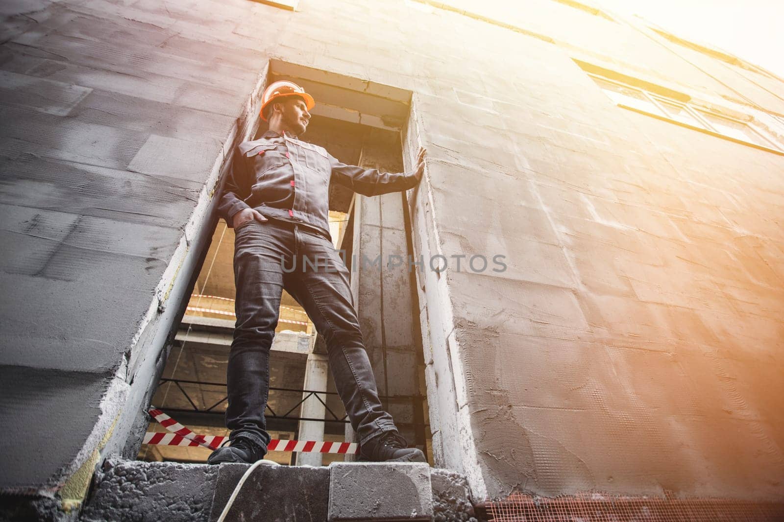 A handyman in overalls and a hard hat stands in the opening of a building under construction.