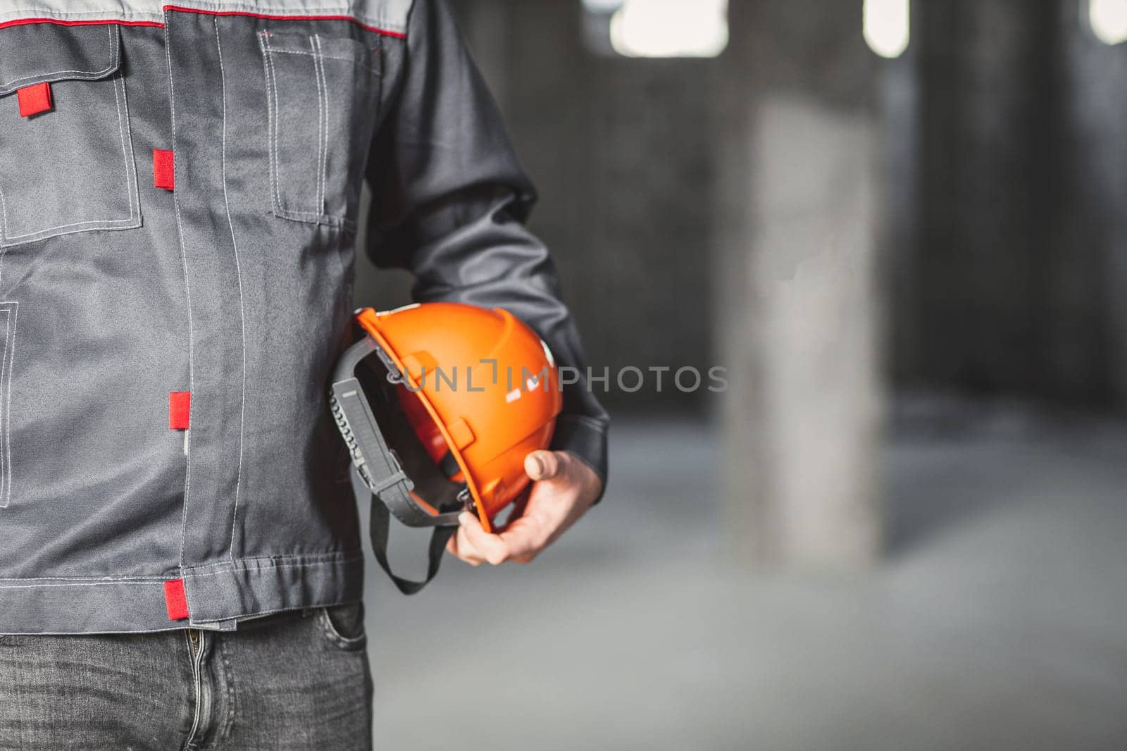 Orange protection helmet is in the hands of a construction worker in overall, copy space, construction site safety concept by Rom4ek