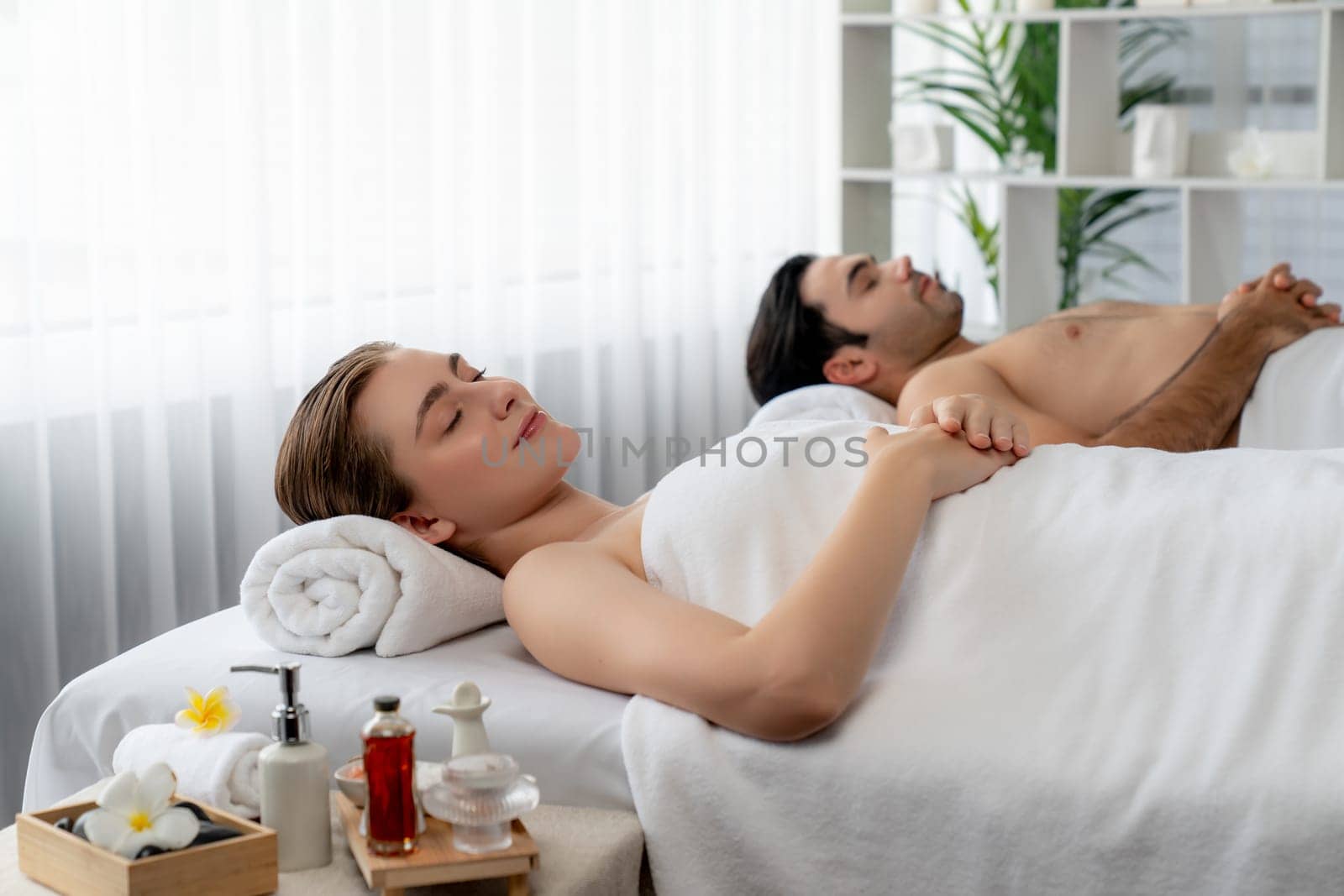 Caucasian couple customer enjoying relaxing anti-stress spa massage and pampering with beauty skin recreation leisure in day light ambient salon spa at luxury resort or hotel. Quiescent