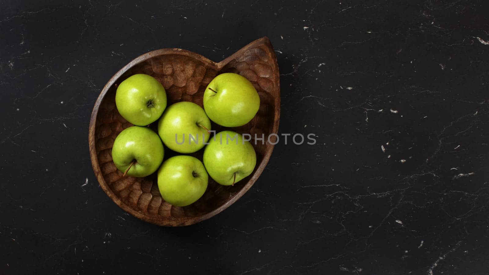 Green apples in wooden carved bowl on black working board, photographed from above. by Ivanko