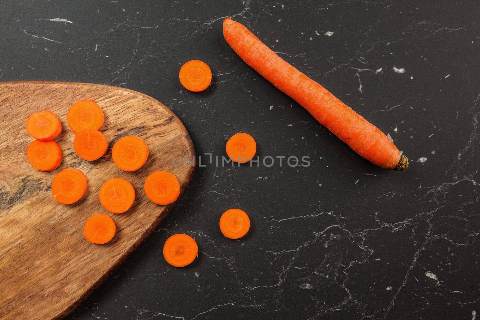 Carrot sliced to small circles and chopping board, whole one next to it on black working desk, photo from above by Ivanko