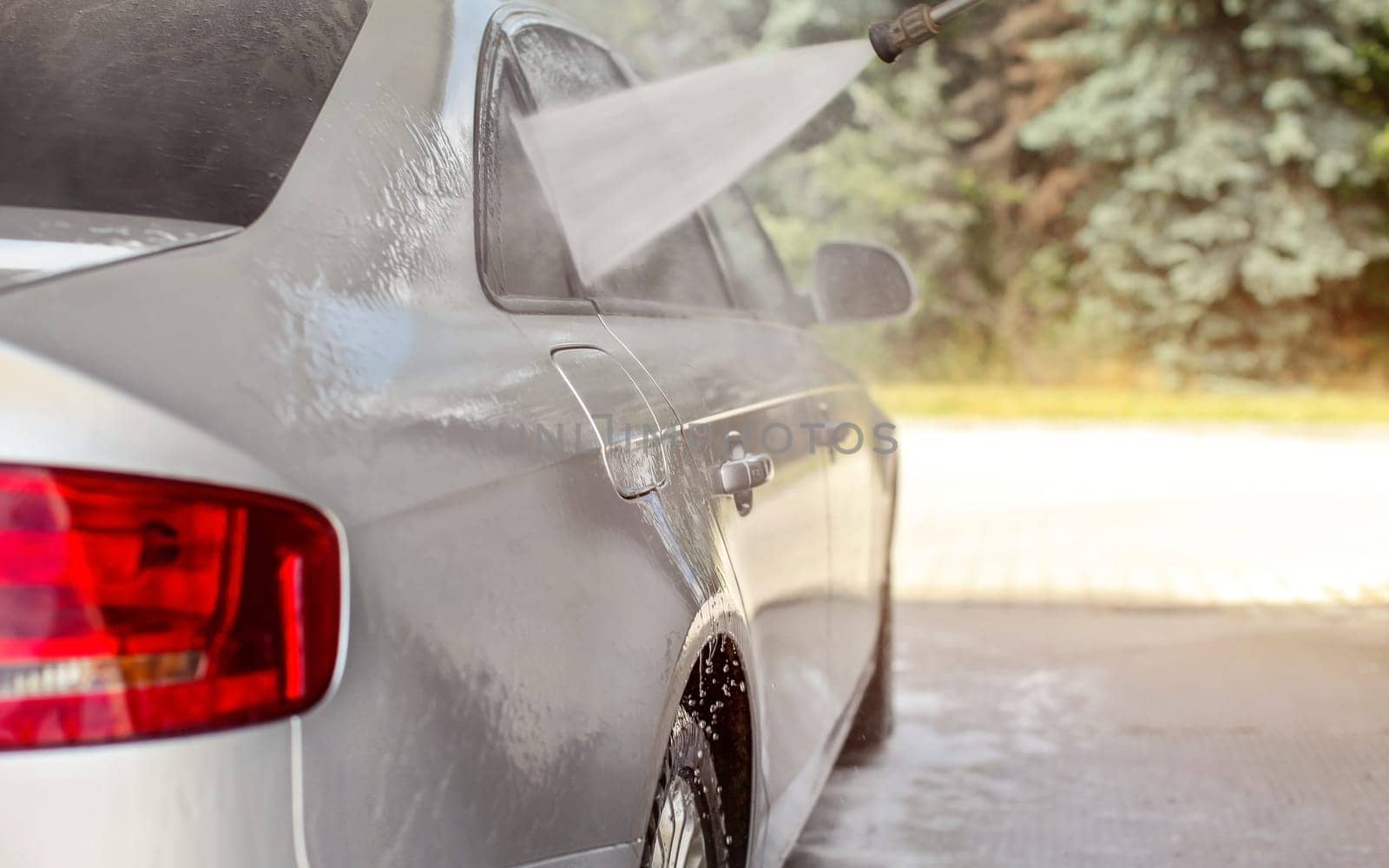 Side of silver car washed in self serve carwash, water spraying from nozzle to window glass.