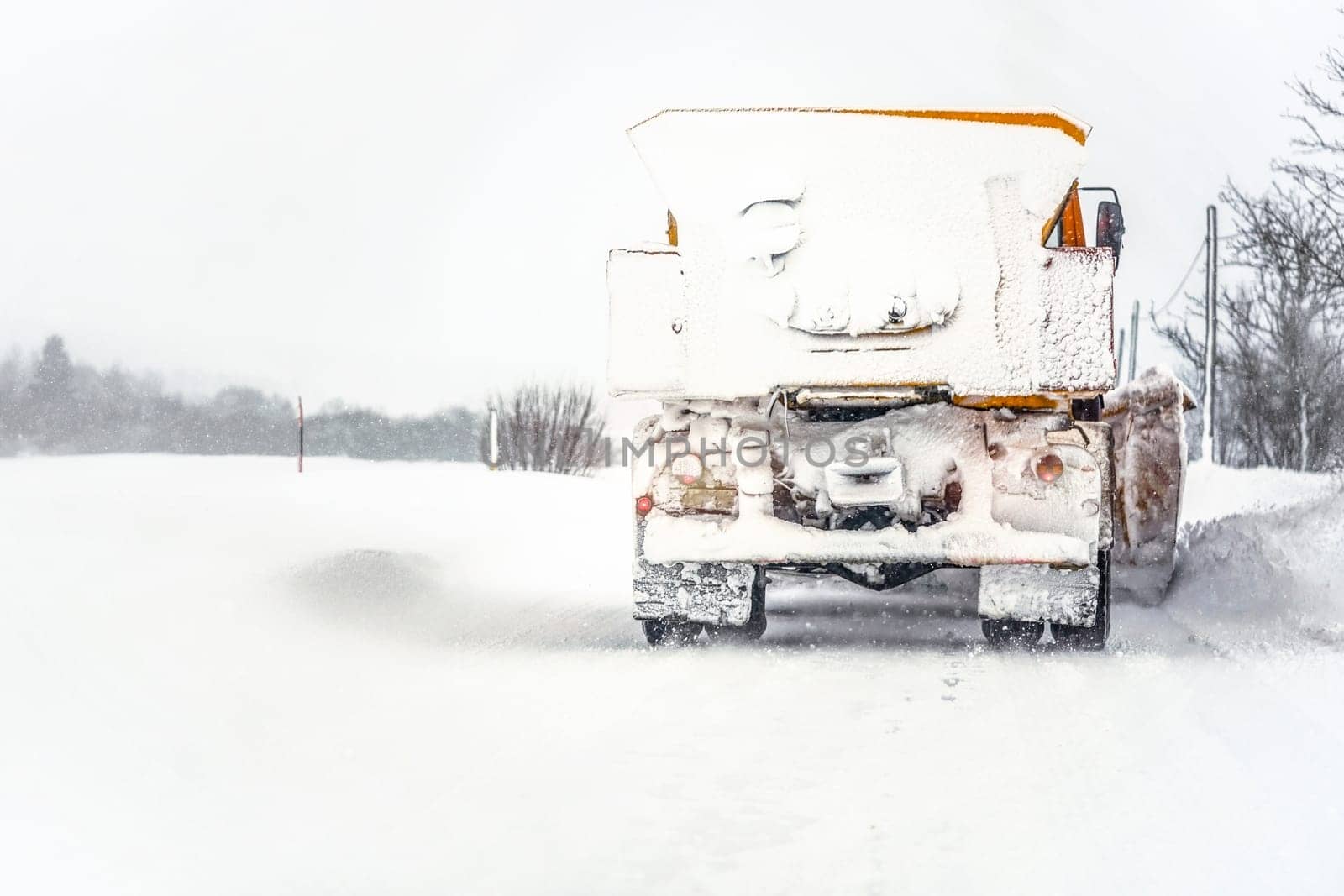 Orange plough truck on path completely covered with snow, gray sky and trees in background, view from back car driving behind - winter road maintenance