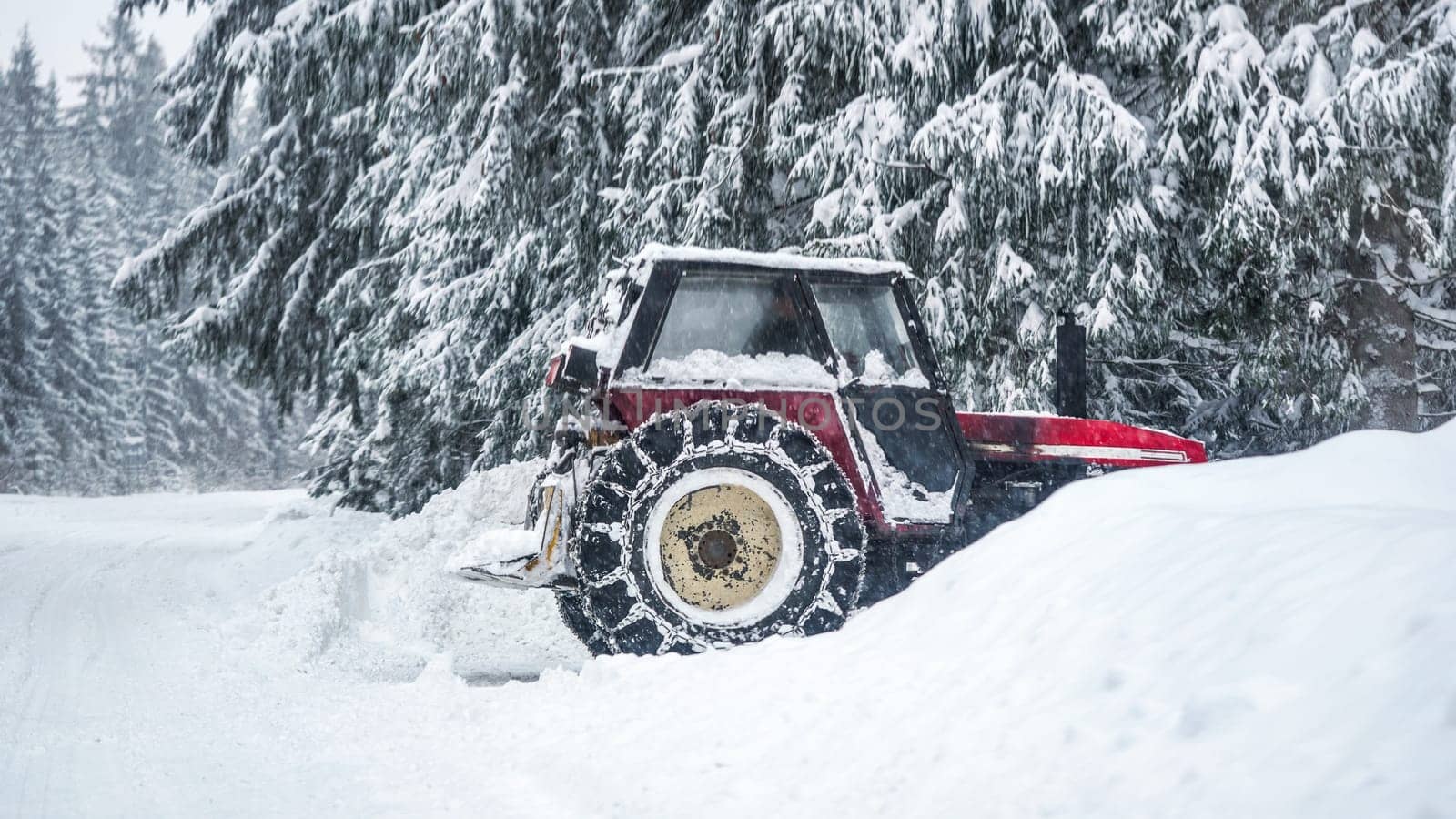 Tractor removing snow from large snowbanks next to forest road, after blizzard in winter by Ivanko