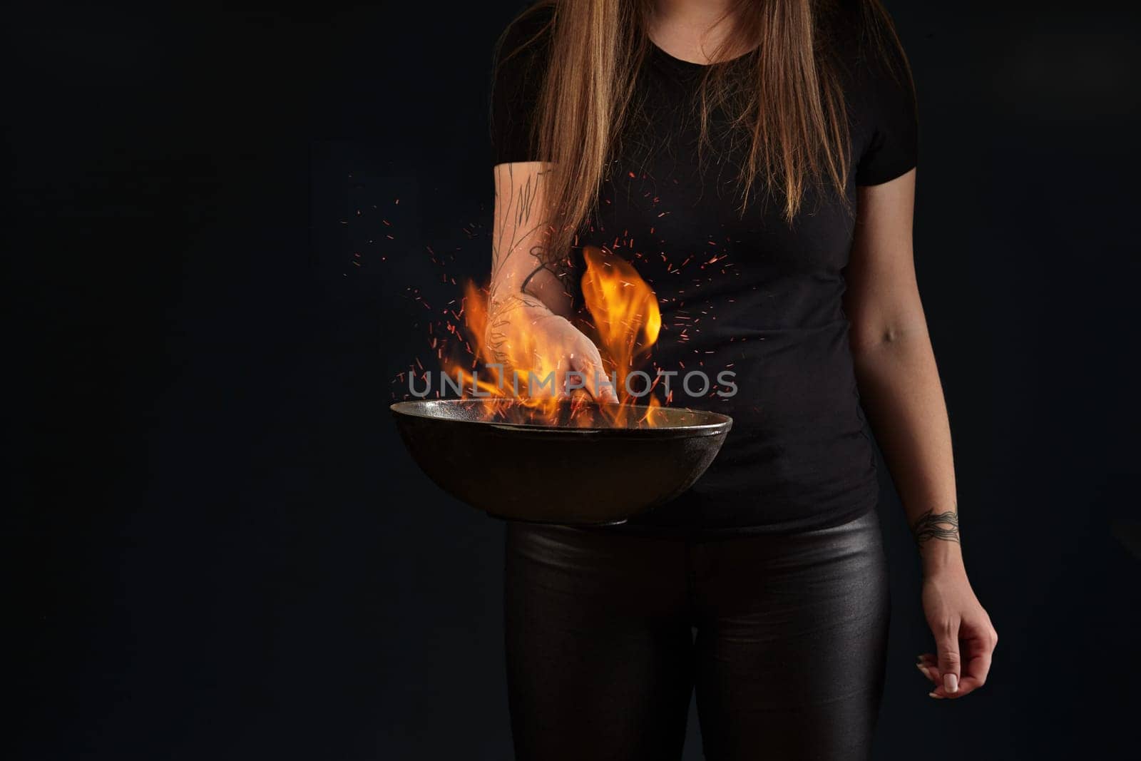 Brunette woman with tattooed hands and long hair, dressed in leather leggings and t-shirt. Holding a wok pan with fire against black studio background. Cooking concept. Close up, copy space
