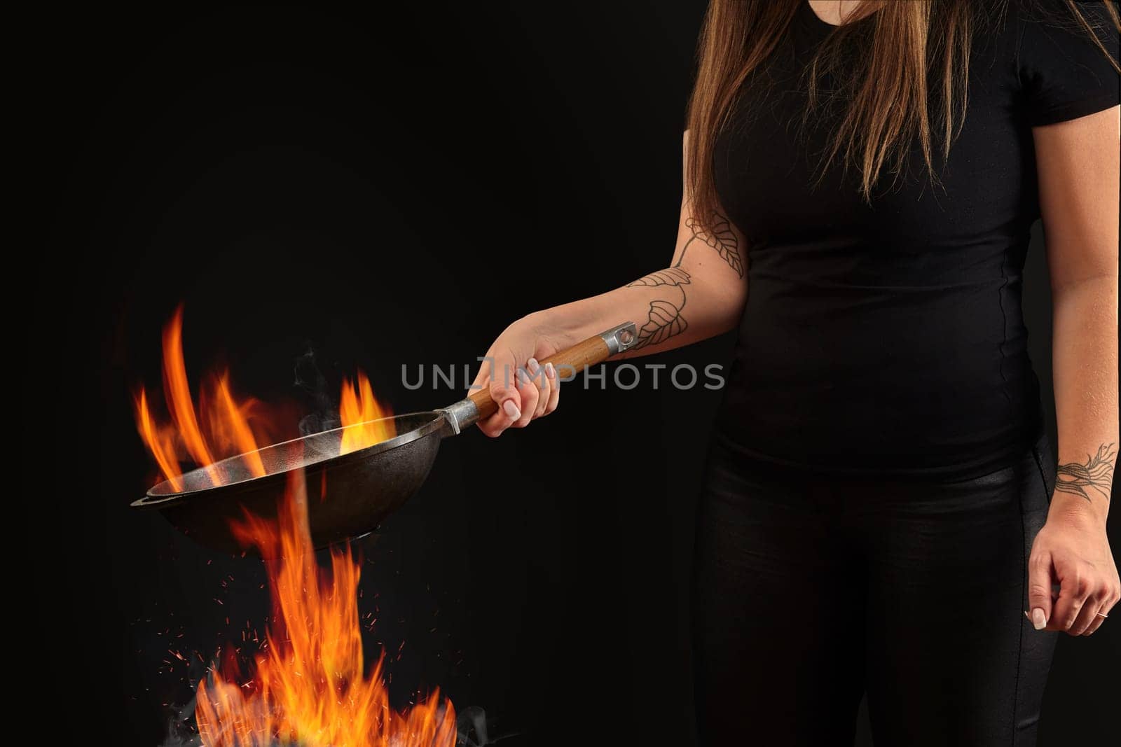 Brunette lady with tattooed hands, dressed in leggings and t-shirt. Holding a wok pan above fire against black background. Cooking concept. Side view by nazarovsergey