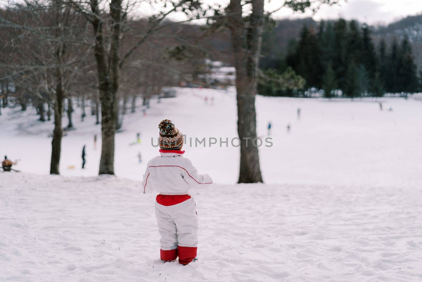 Little girl stands on a snow-covered hill and looks at the skating rink with lugers and skiers below. Back view. High quality photo