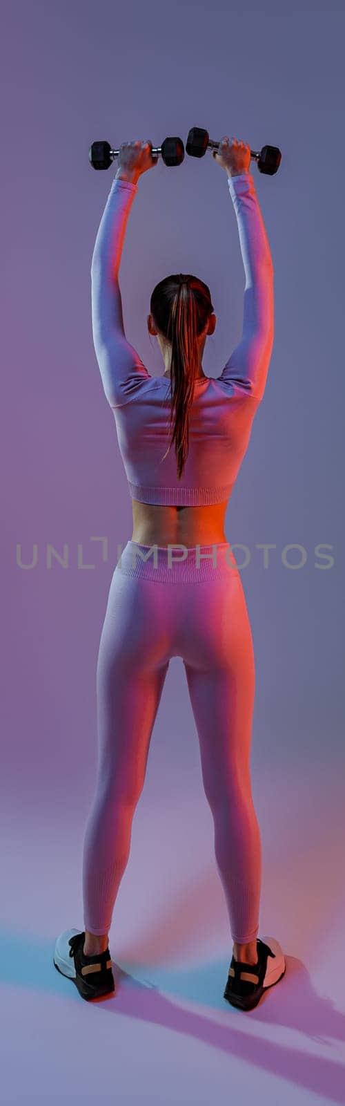 Back view of fitness woman doing exercises with dumbbells on studio background with colored filter by Yaroslav_astakhov
